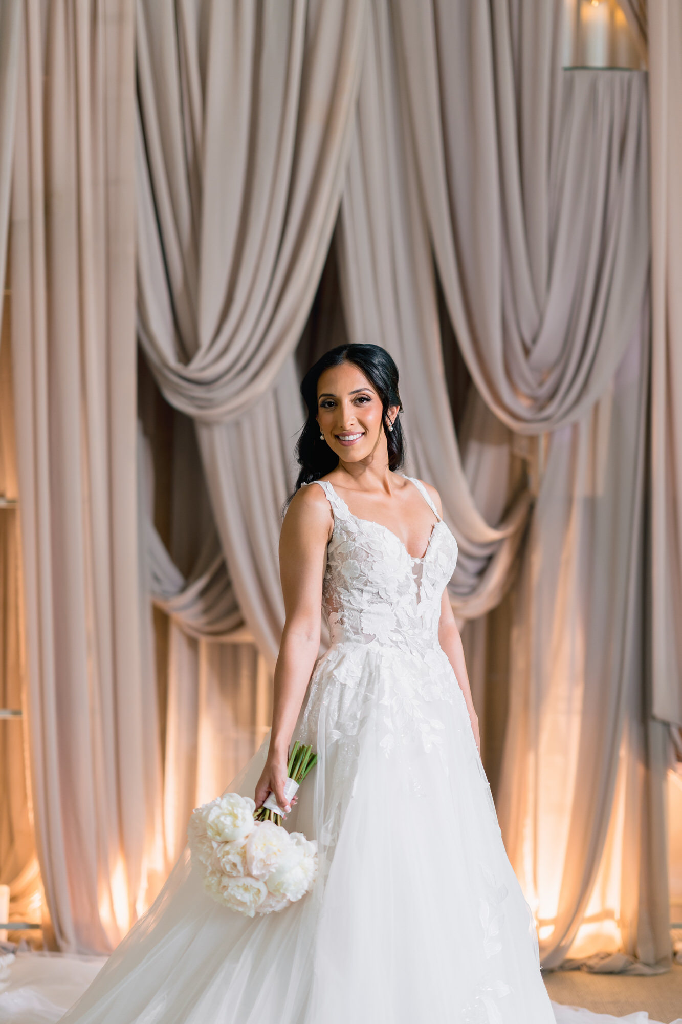 The bride standing with a white bouquet in front of draped curtains and soft candlelight