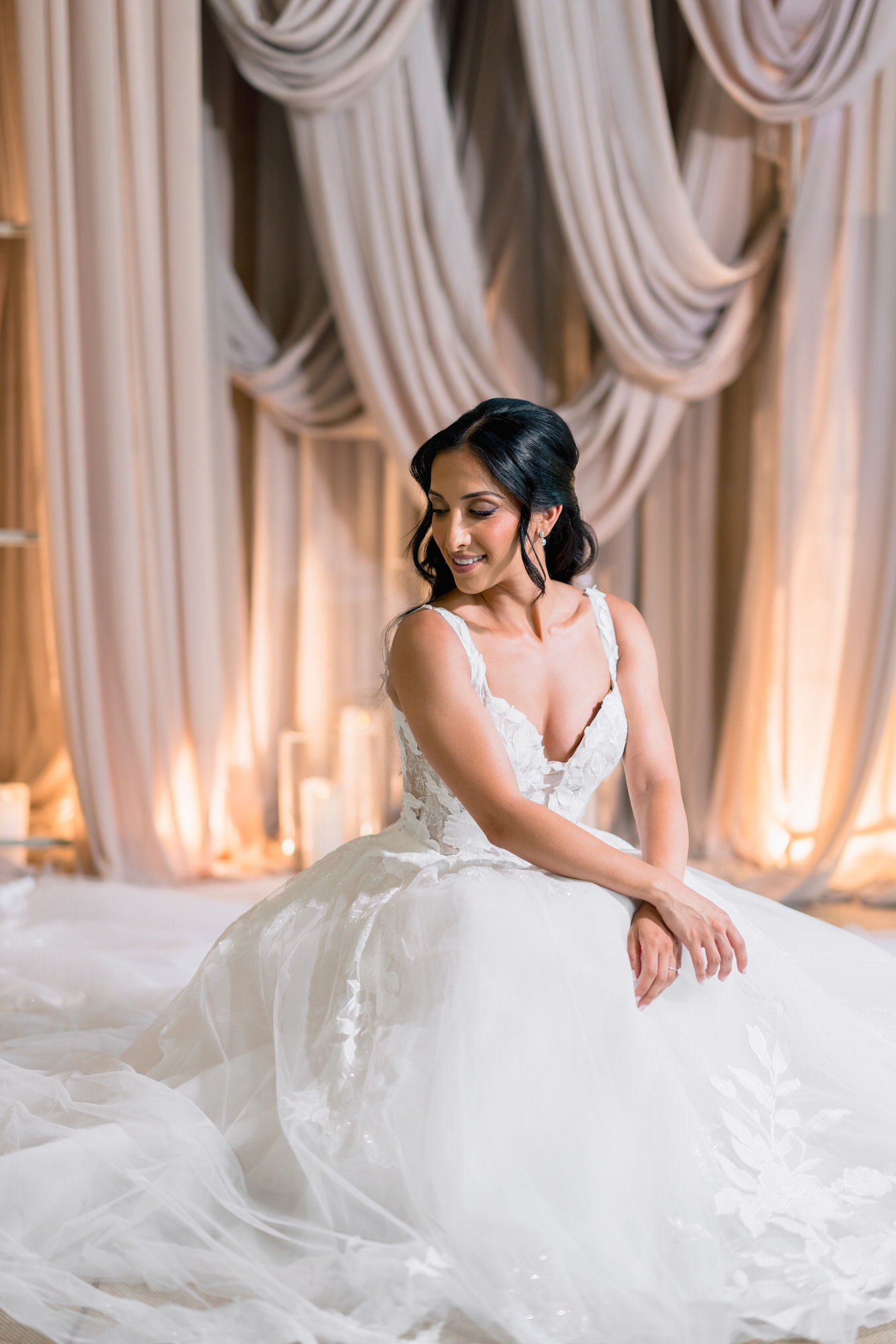 The bride seated on the floor in her wedding gown before draped taupe curtains lit by candles