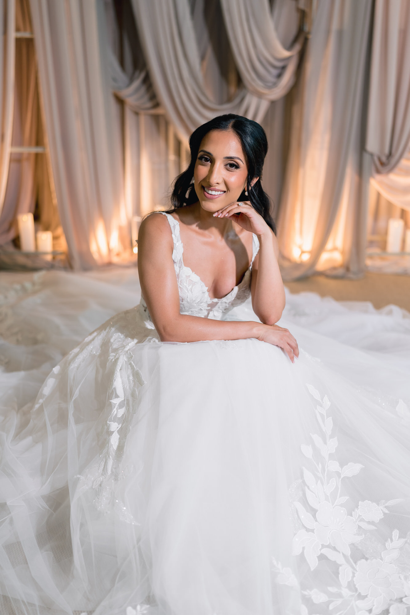 The bride seated on the floor, smiling, in a gown with lace appliqué on the tulle skirt