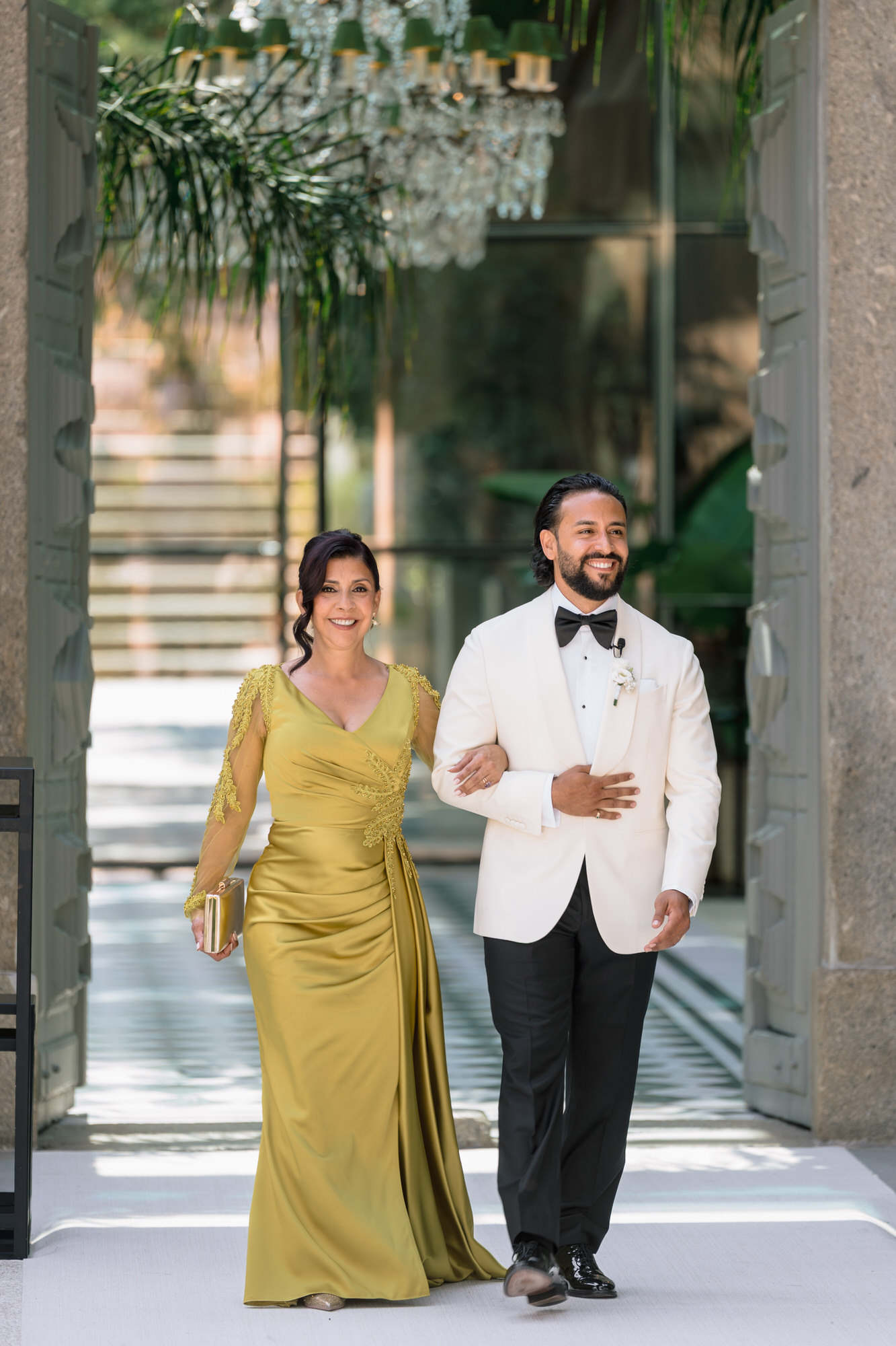 The groom in a white dinner jacket walking with a woman in a mustard-yellow satin gown beneath a stone arch