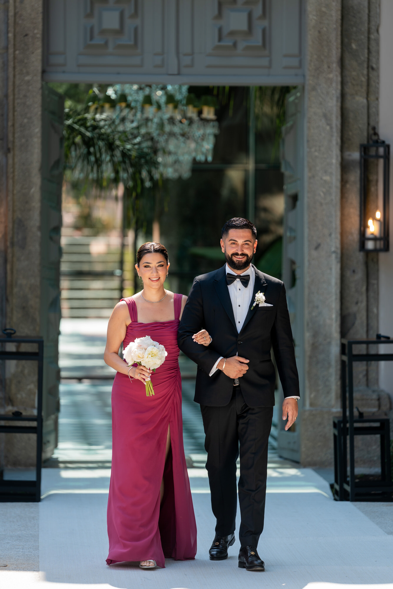 A bridesmaid in a berry gown walking arm in arm with a groomsman in a black suit, white bouquet in hand