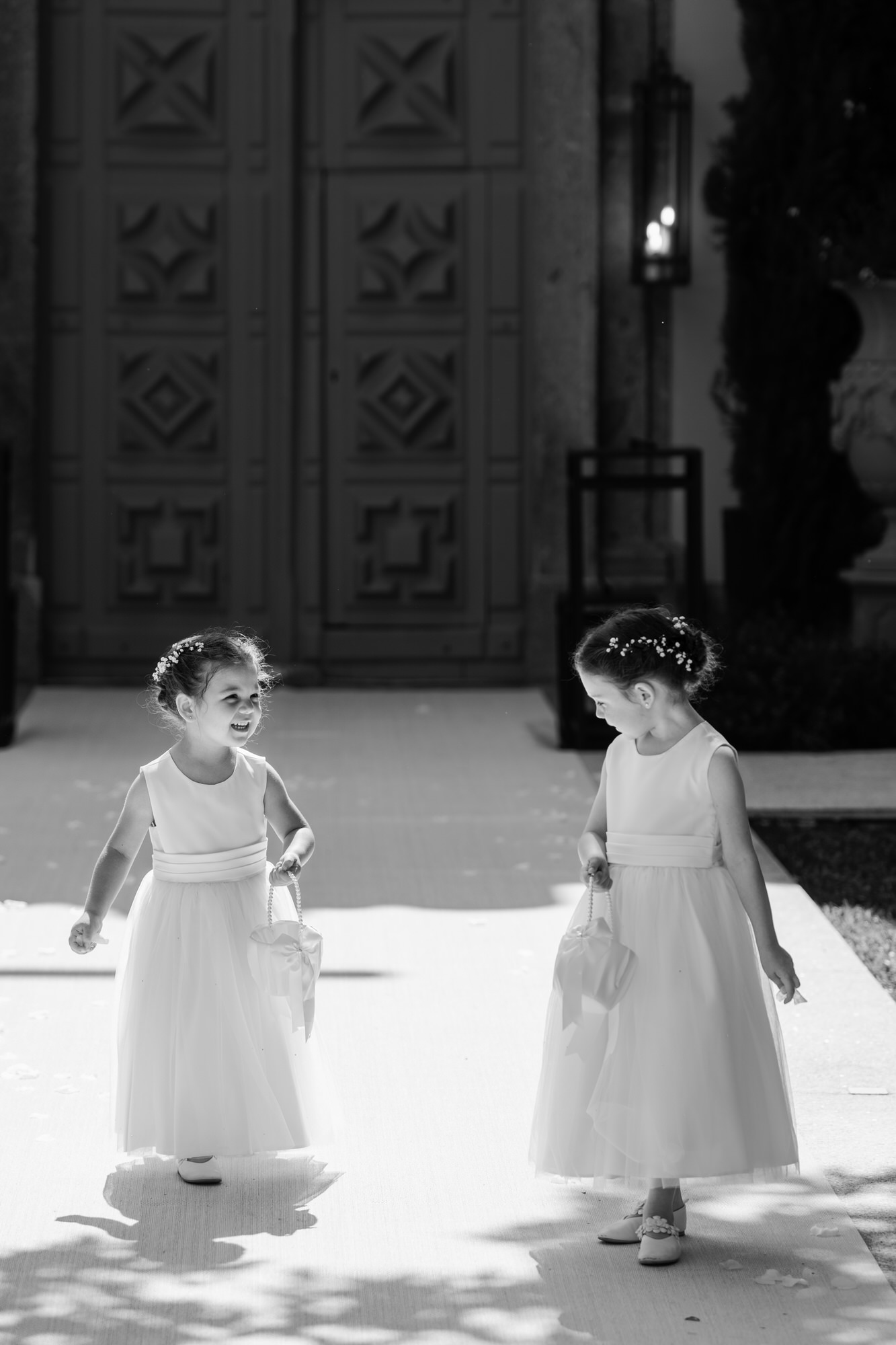 Black-and-white image of two flower girls in white dresses on a sunlit path before dark wooden doors