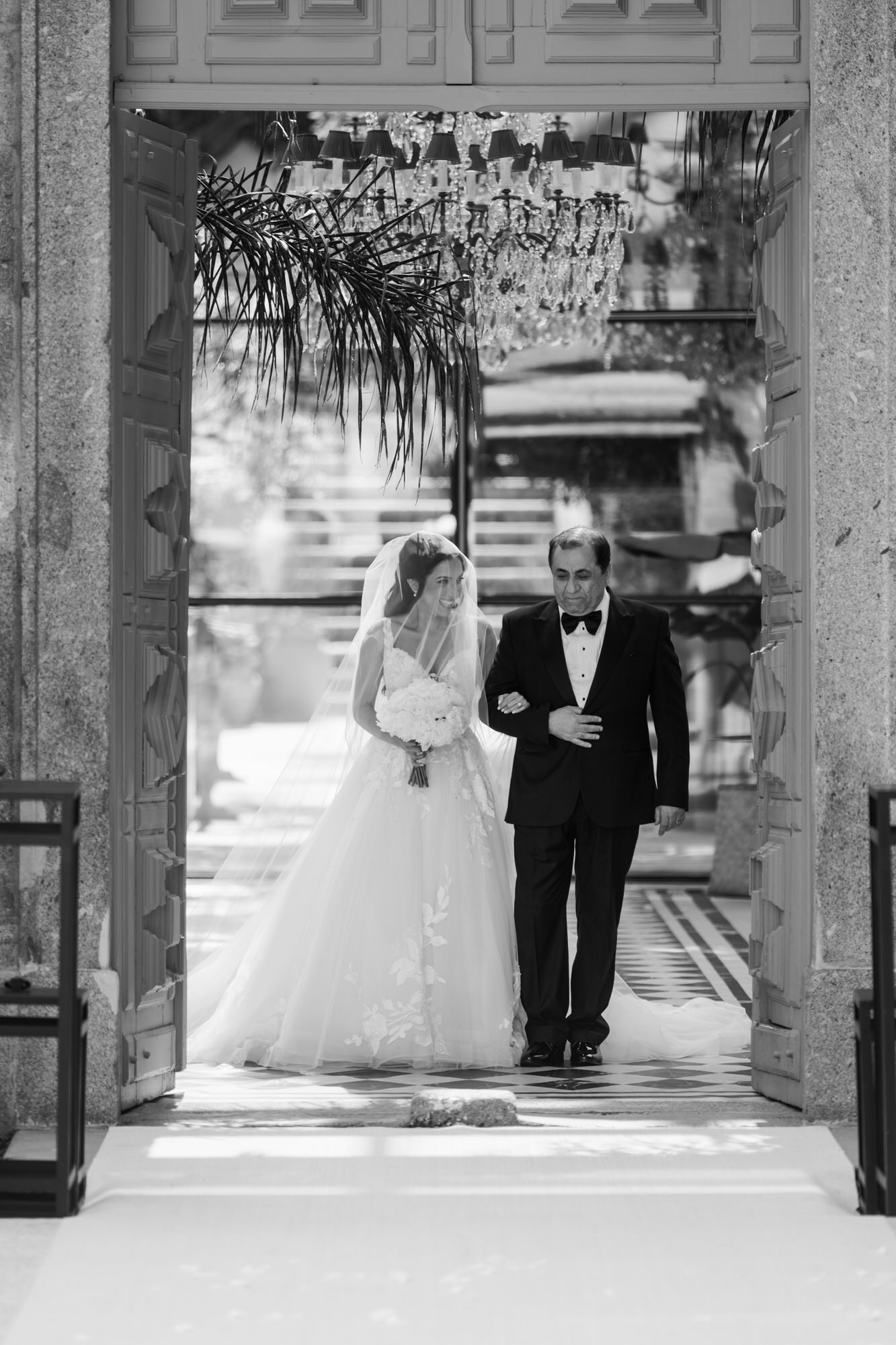 Black-and-white image of the bride and an older man walking through a stone archway under a hanging chandelier