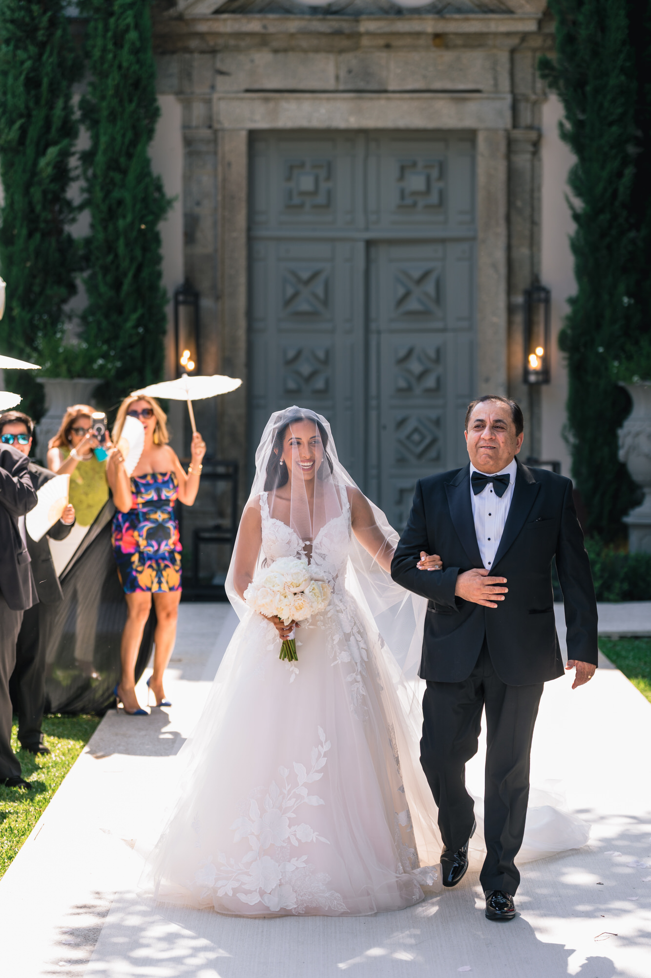 The bride in a long veil walking down the aisle with a man in a black suit, guests smiling beside them