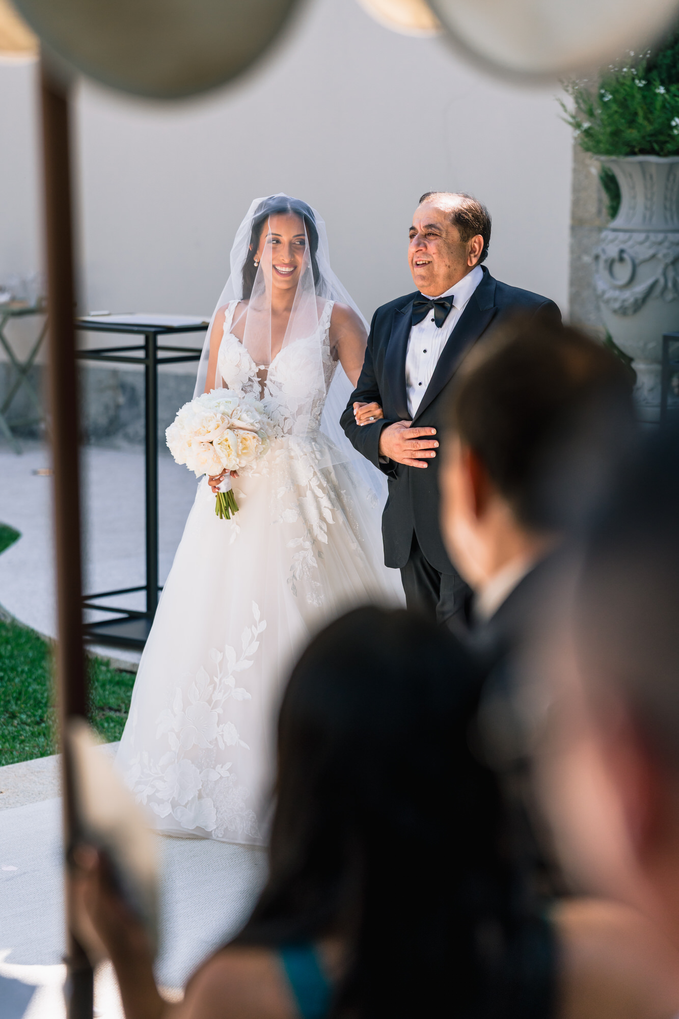 The bride in her veil walking with an older man in a dark suit as guests look on