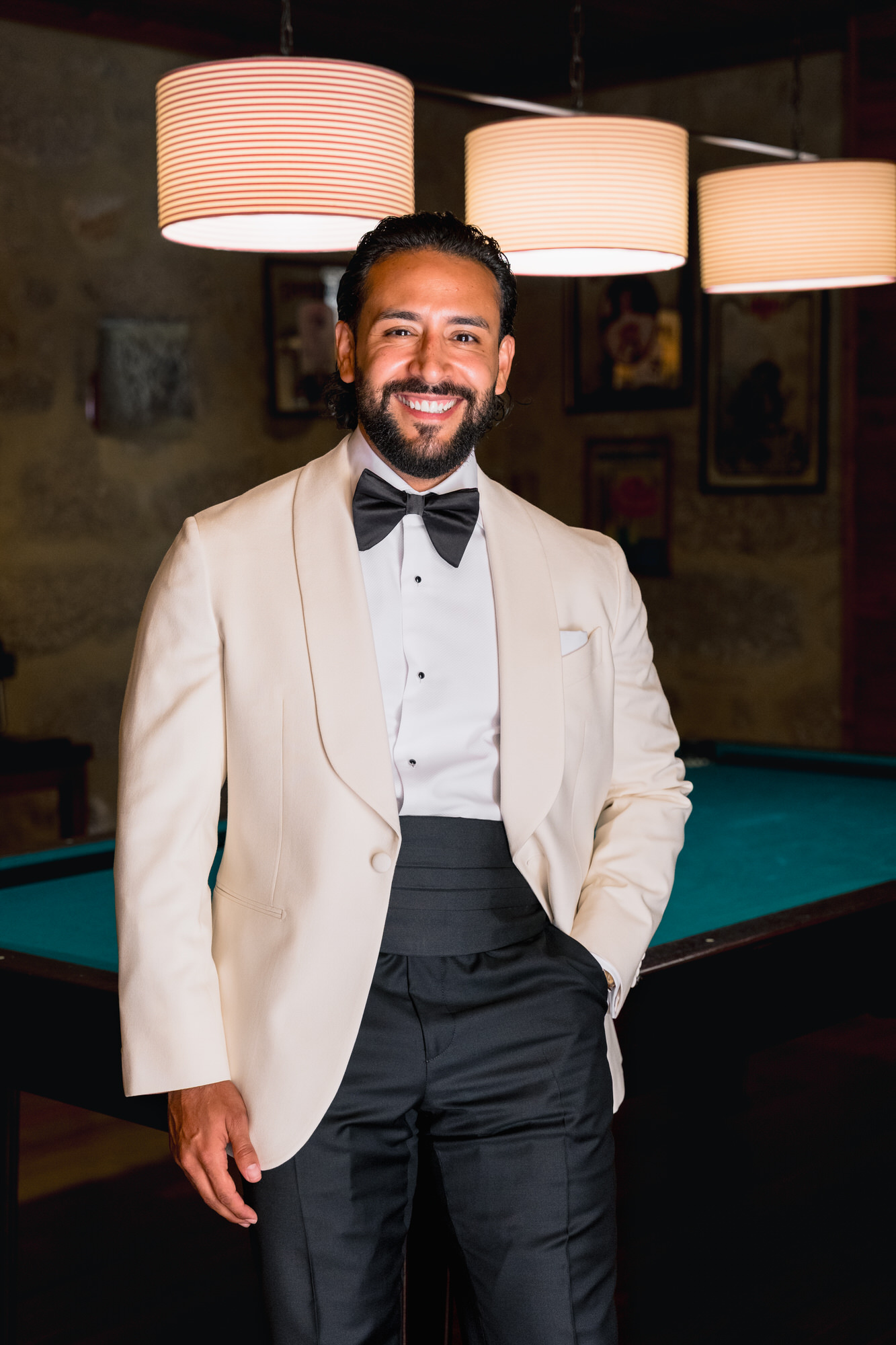 The groom smiling in a cream dinner jacket and black bow tie, beside a pool table under warm pendant lamps