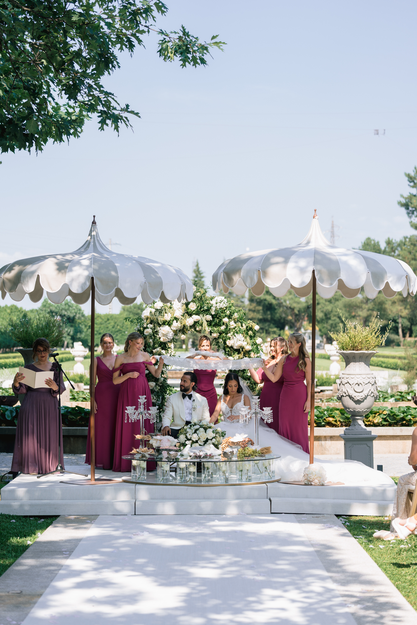 The ceremony table covered in flowers beneath white scalloped parasols, guests in pink and berry dresses behind