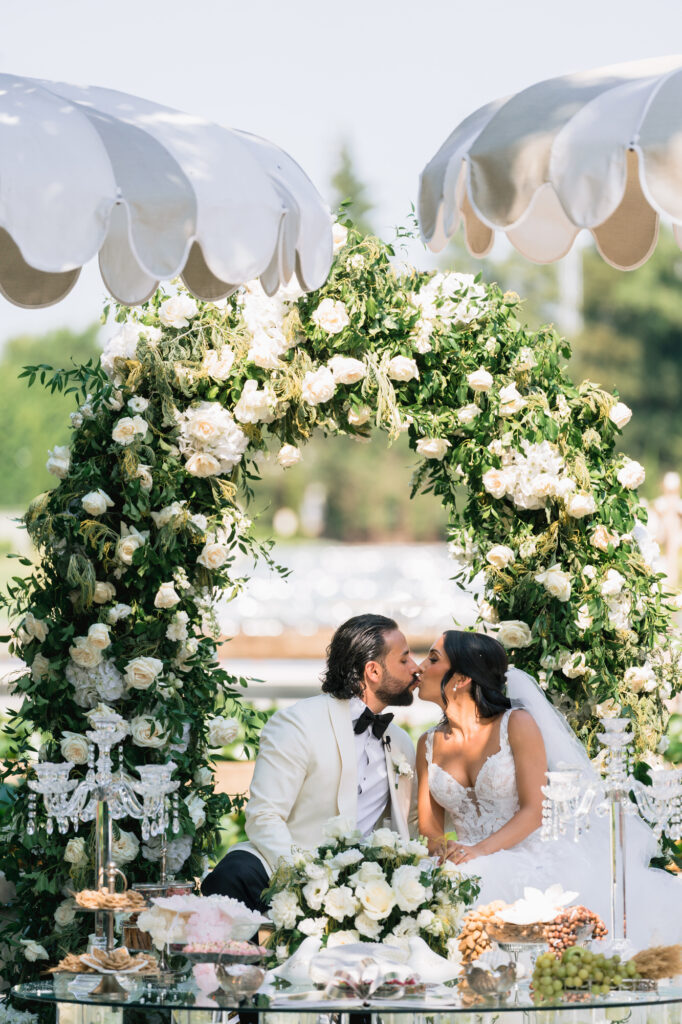 Bride and groom kiss under a white floral arch at an outdoor wedding, with a dessert table in front.",