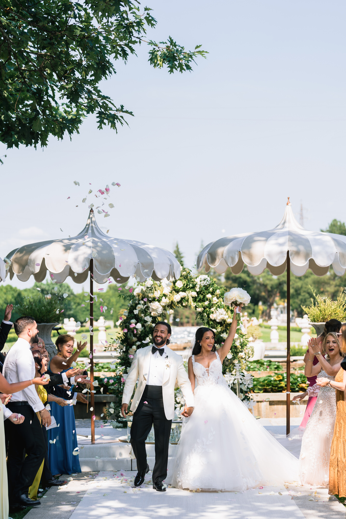 The couple walking between guests under white parasols, a floral arch behind them