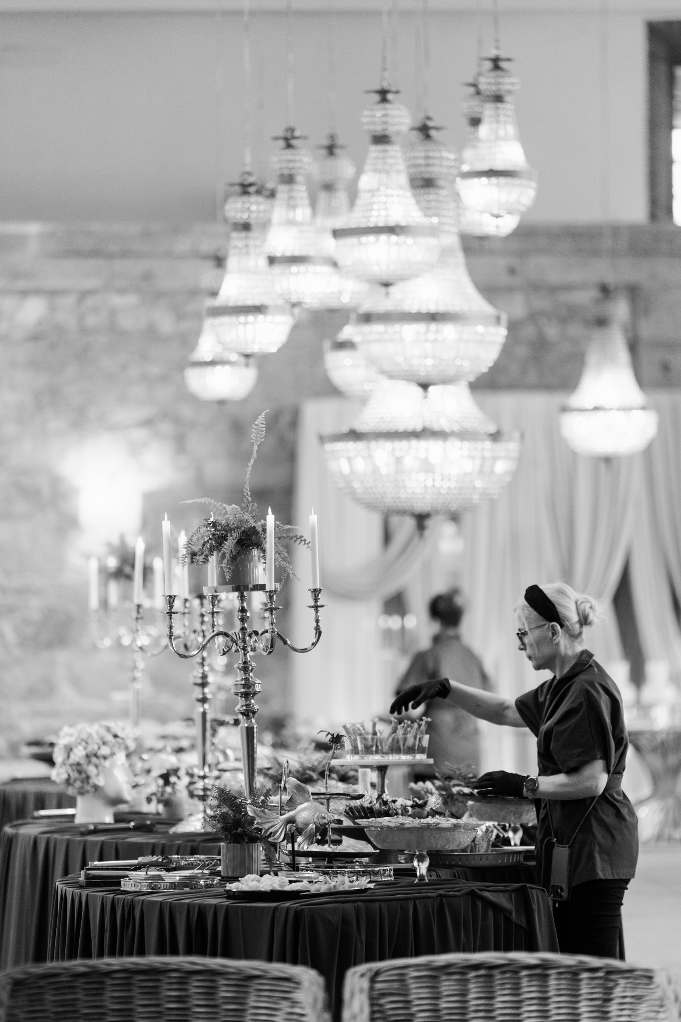 Black-and-white image of the reception hall with tiered chandeliers and round tables being prepared