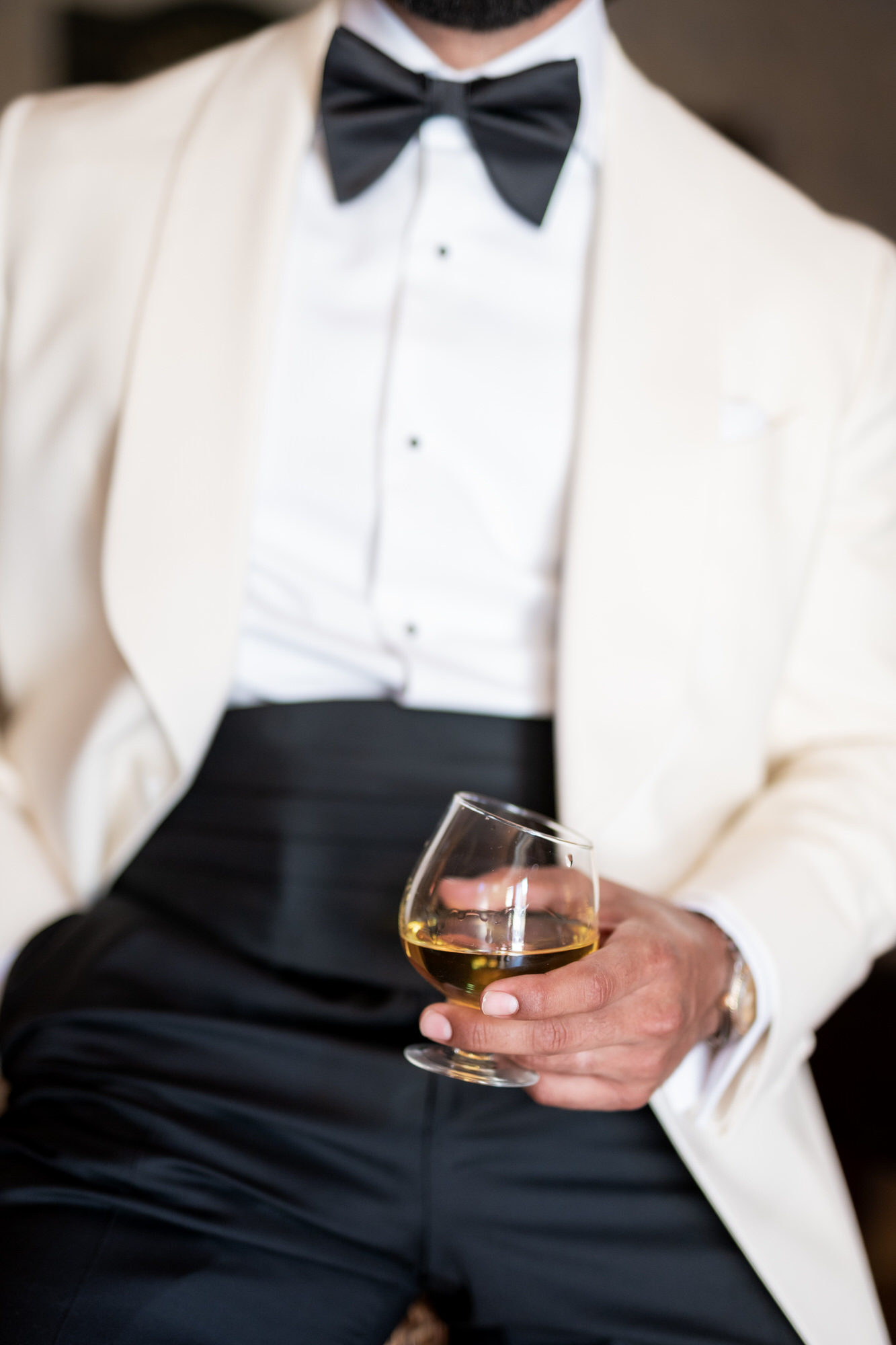 Close-up of the groom in a white dinner jacket and black bow tie, holding a glass of whisky at waist level