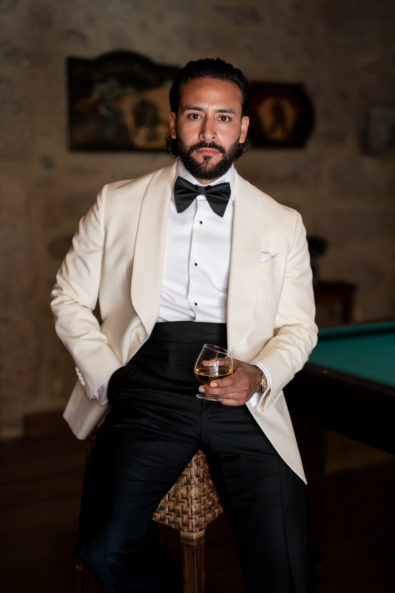 The groom leaning against a pool table in a cream dinner jacket, whisky glass in hand, looking into the camera