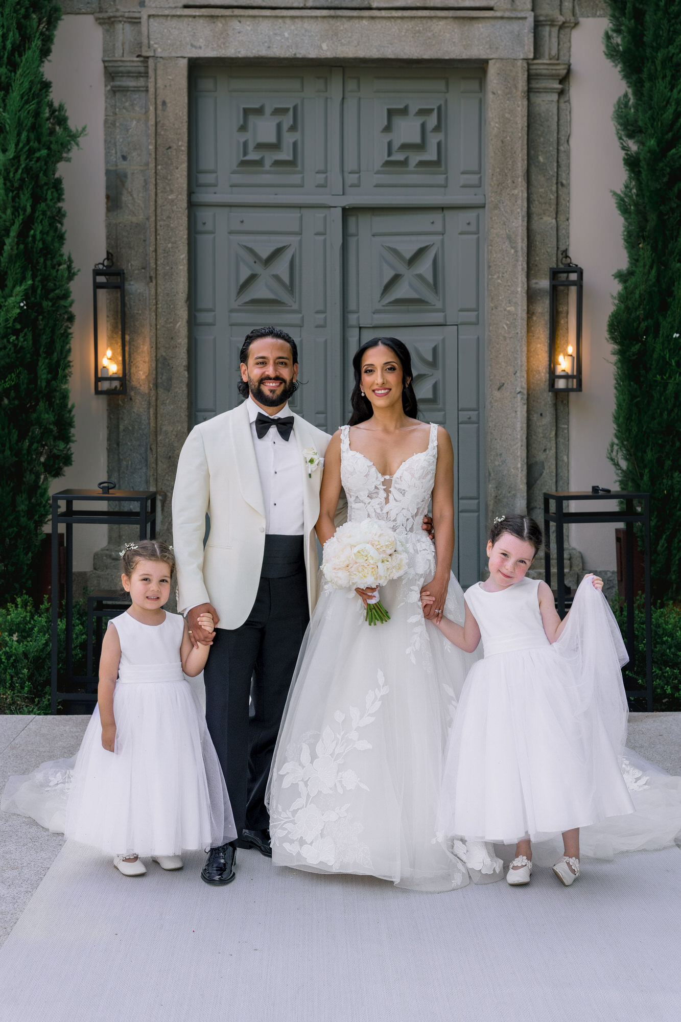 Hana and Andres posing with two flower girls in white dresses in front of the stone doors