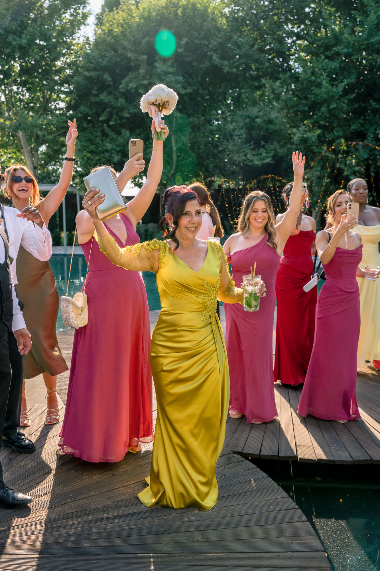 Female guests in berry, pink and yellow dresses raising their bouquets on a wooden deck by the water