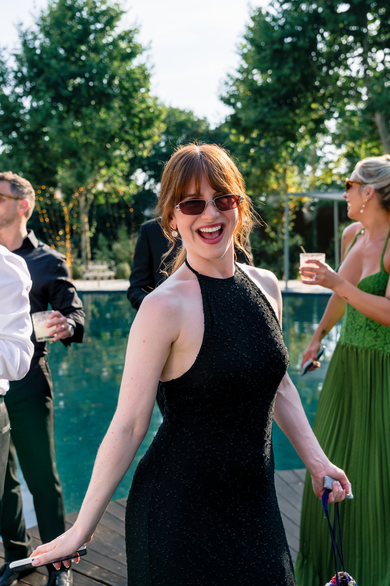 A guest in a black dress smiling beside the pool, friends gathered behind her