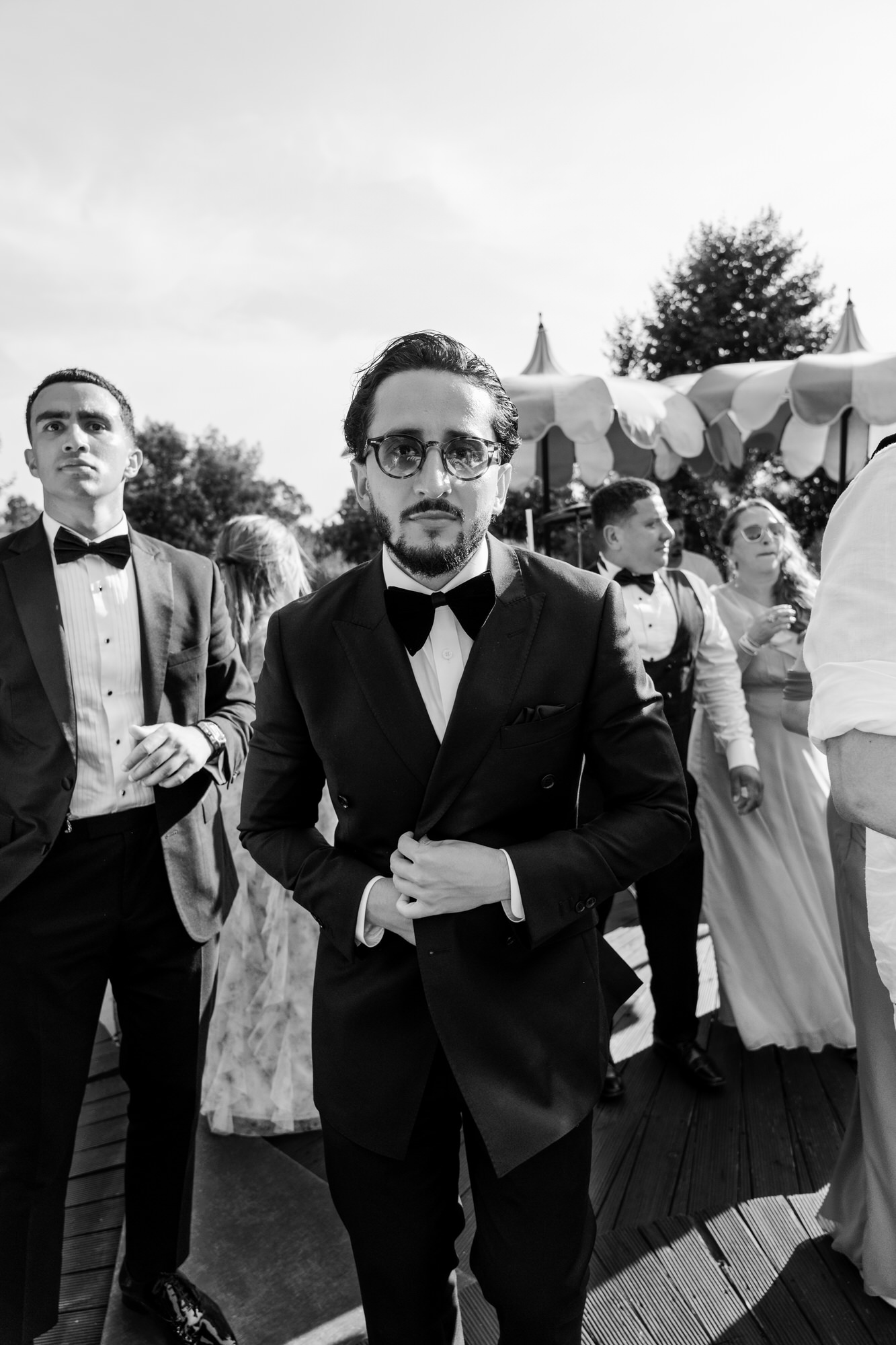 Black-and-white image of a guest in a tuxedo and glasses laughing among other guests