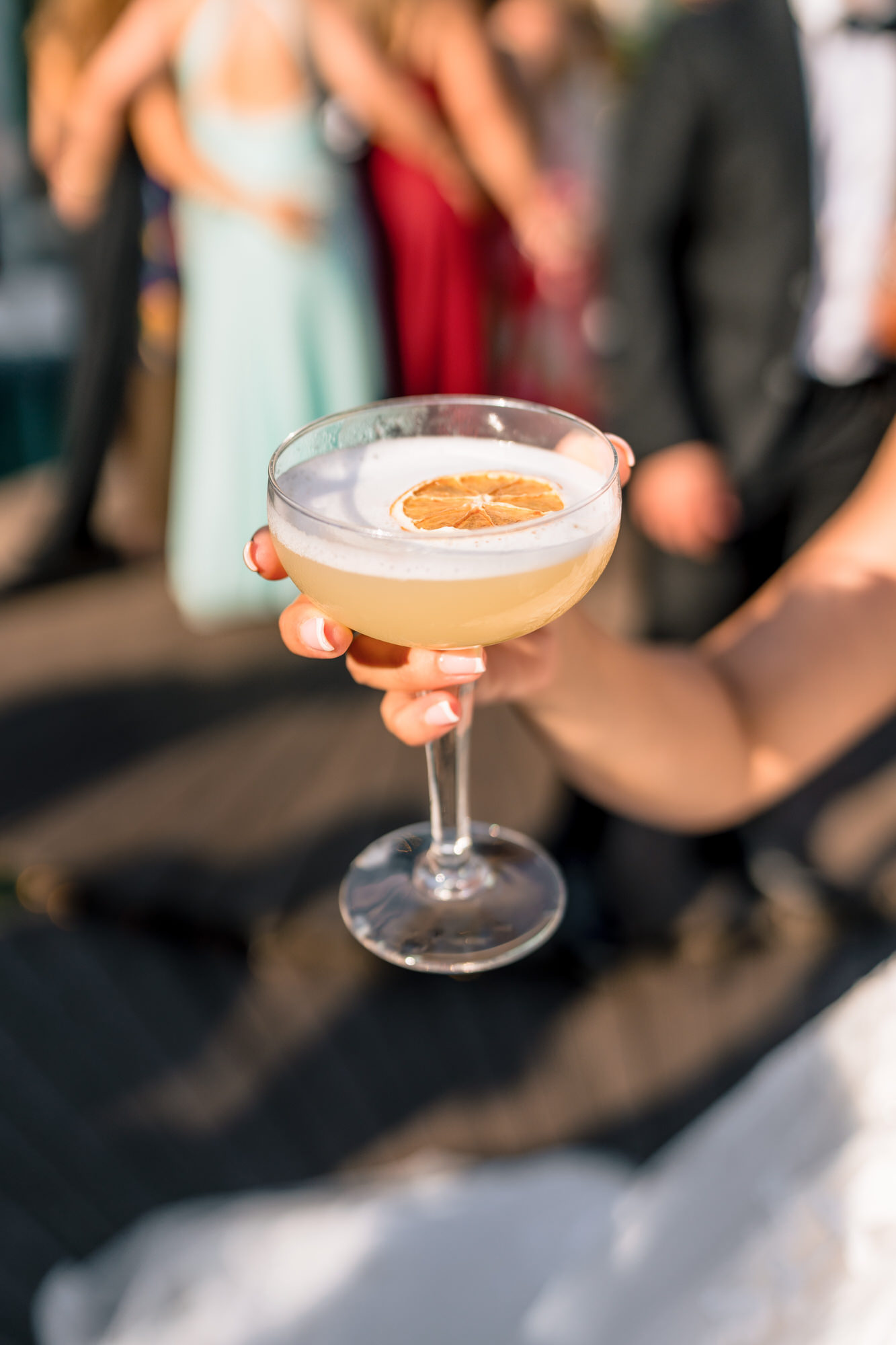 A hand holding a cocktail coupe, with guests blurred in the background