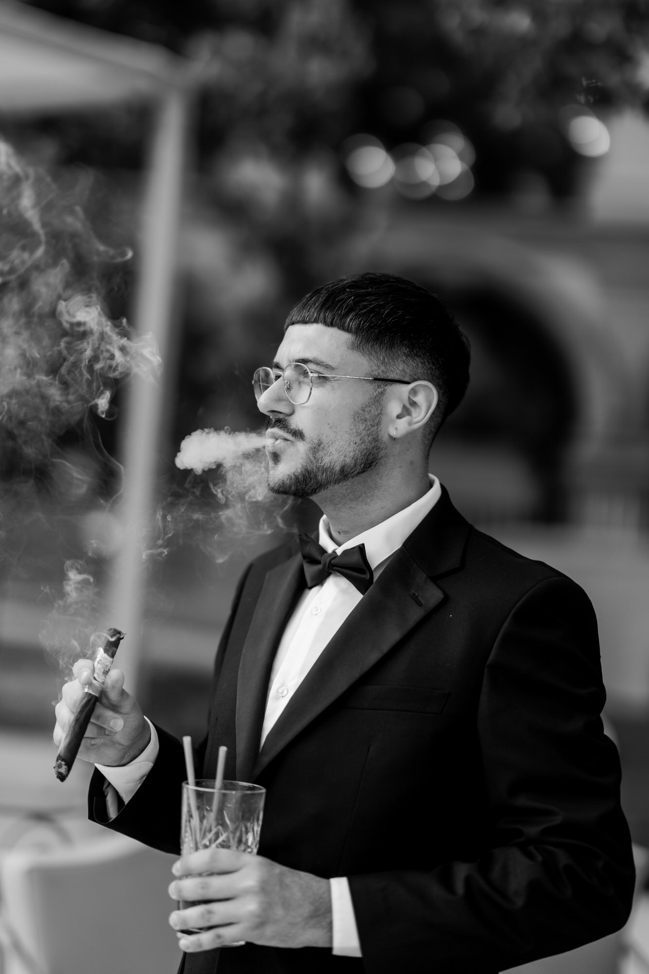Black-and-white portrait of a guest in a tuxedo, bow tie and glasses, holding a drink
