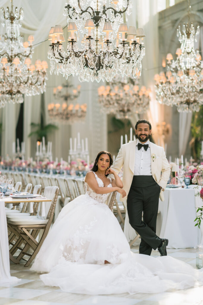 Bride in a white wedding dress sits beside a groom in a white tuxedo and black bow tie at an elegant, chandeliers-lit reception hall.