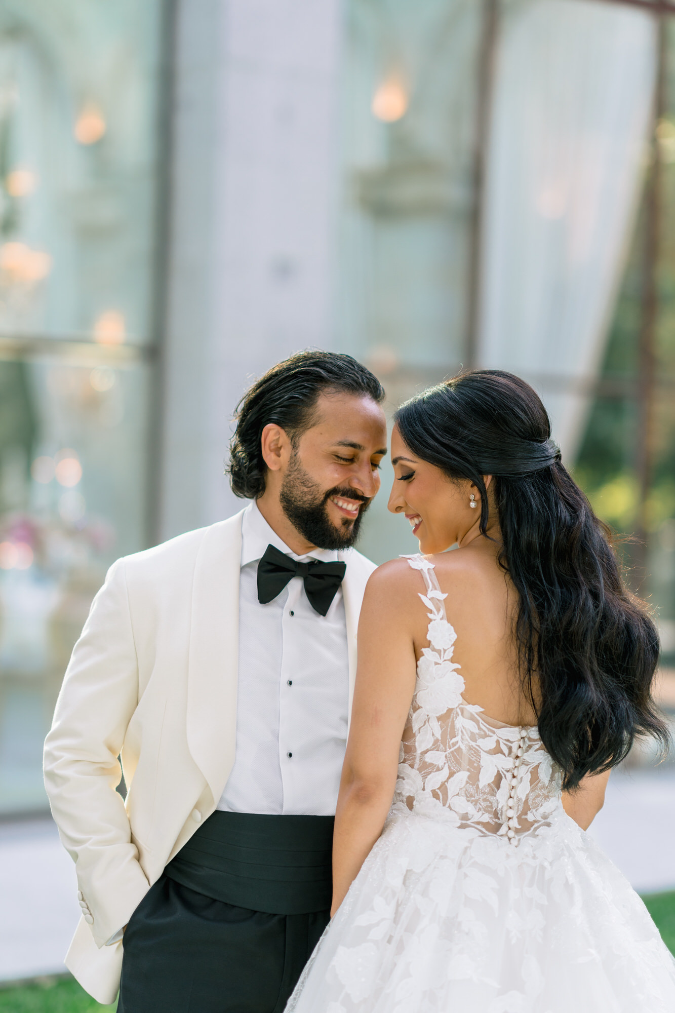 Hana and Andres foreheads together, she in a sheer lace bodice and he in a white dinner jacket, beside glass doors