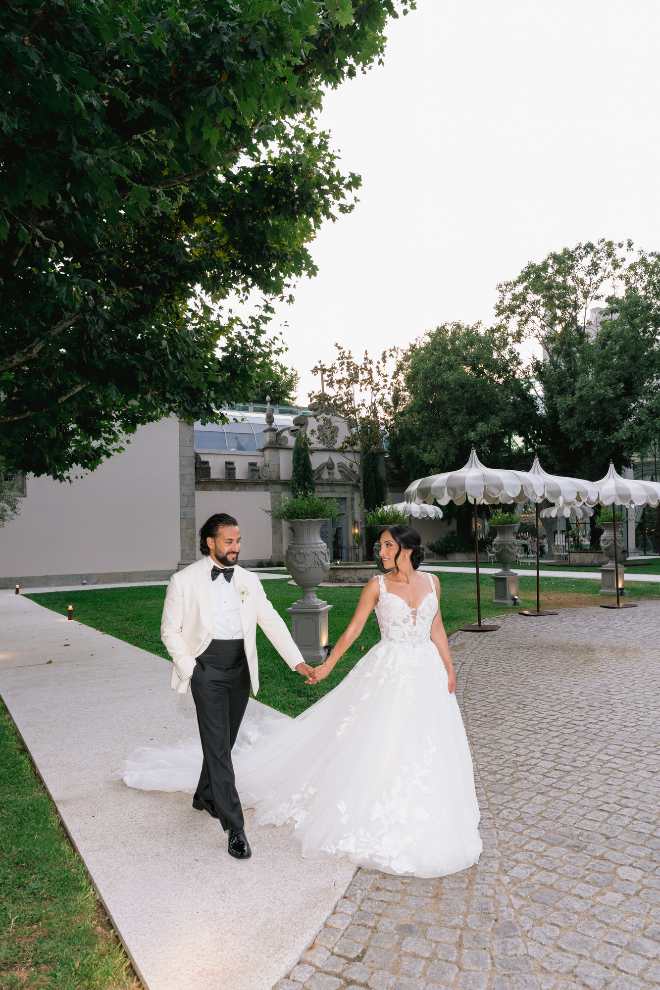 The couple walking hand in hand along a brick path through the gardens in warm light