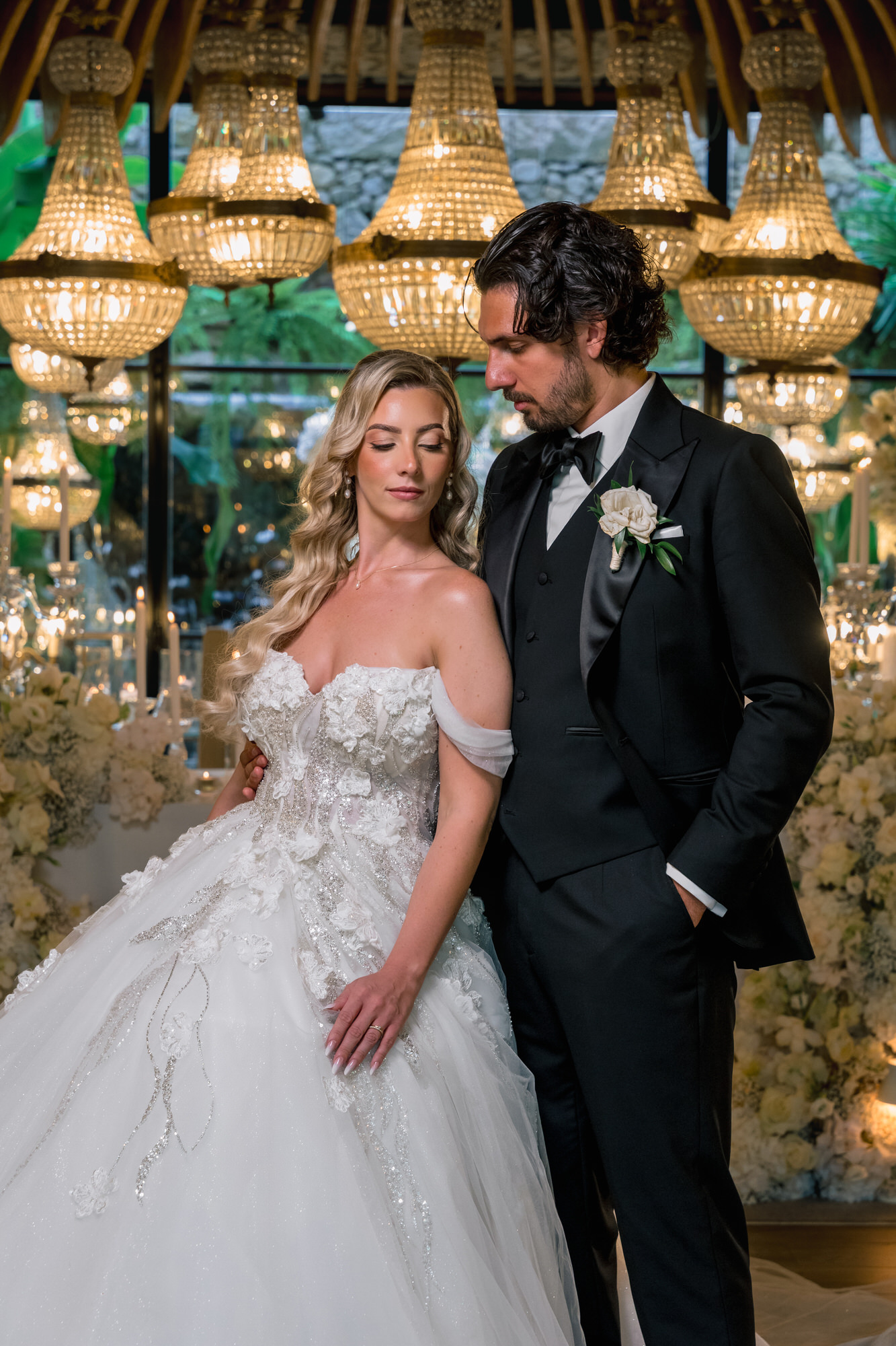 The couple in the reception hall, the bride in a tulle gown and the groom in a black tuxedo, beneath golden pendant lamps