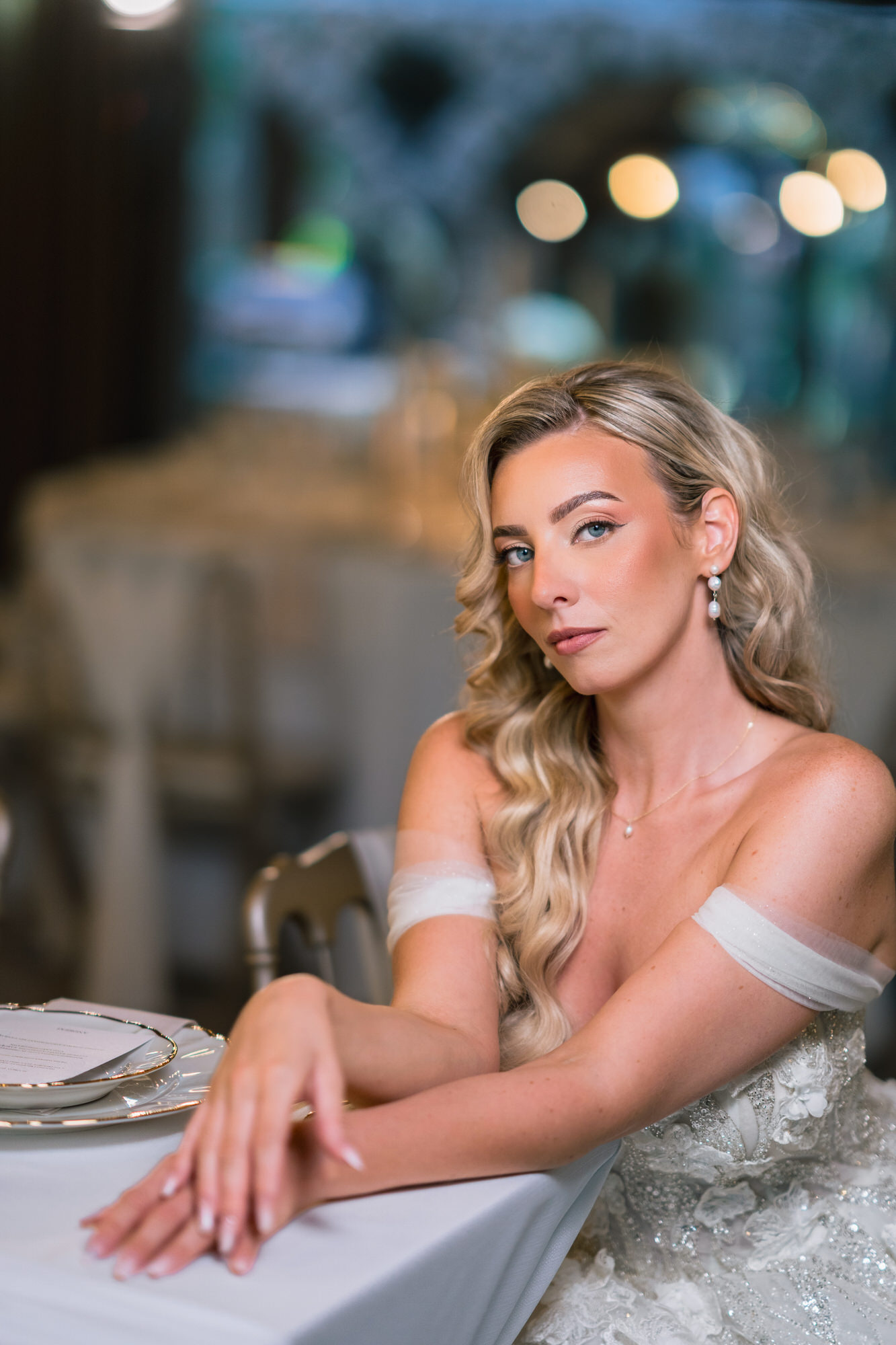 The bride seated at a table, her arms crossed and gaze on the camera, in an off-the-shoulder gown
