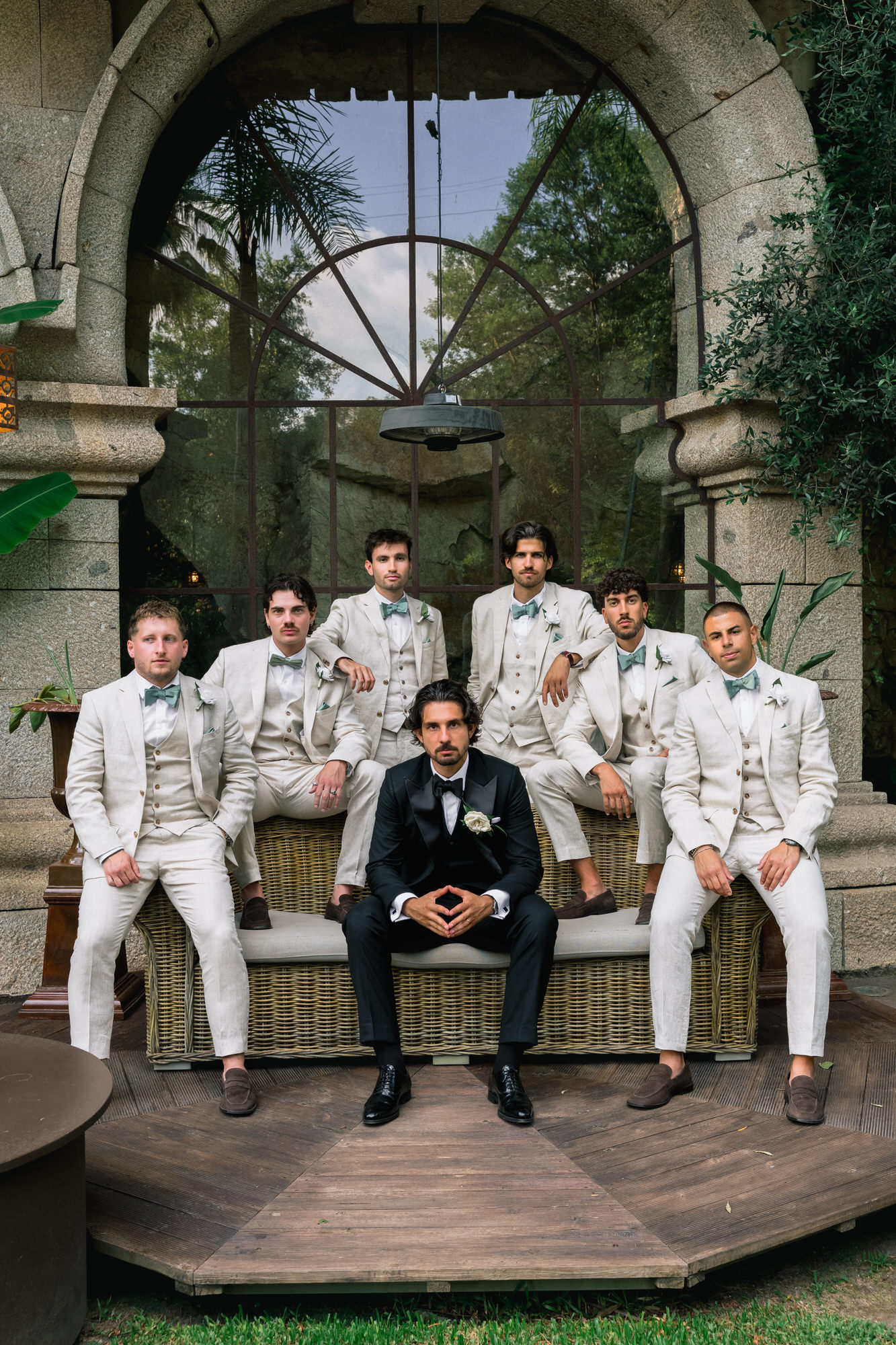 The groom seated among his groomsmen in cream suits under a stone arch