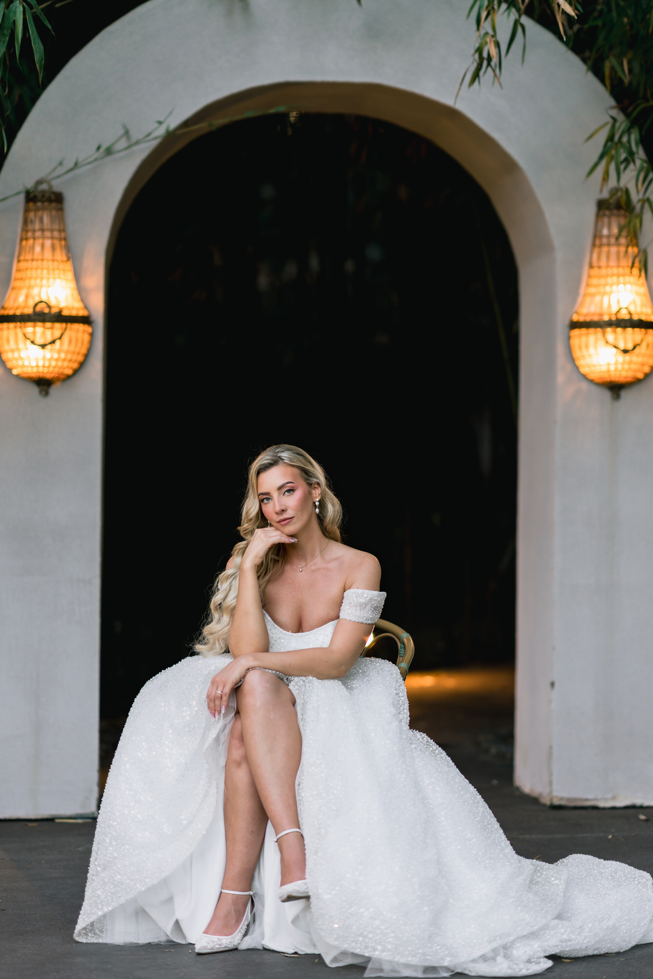 The bride seated in her white gown in front of an arched doorway flanked by lanterns