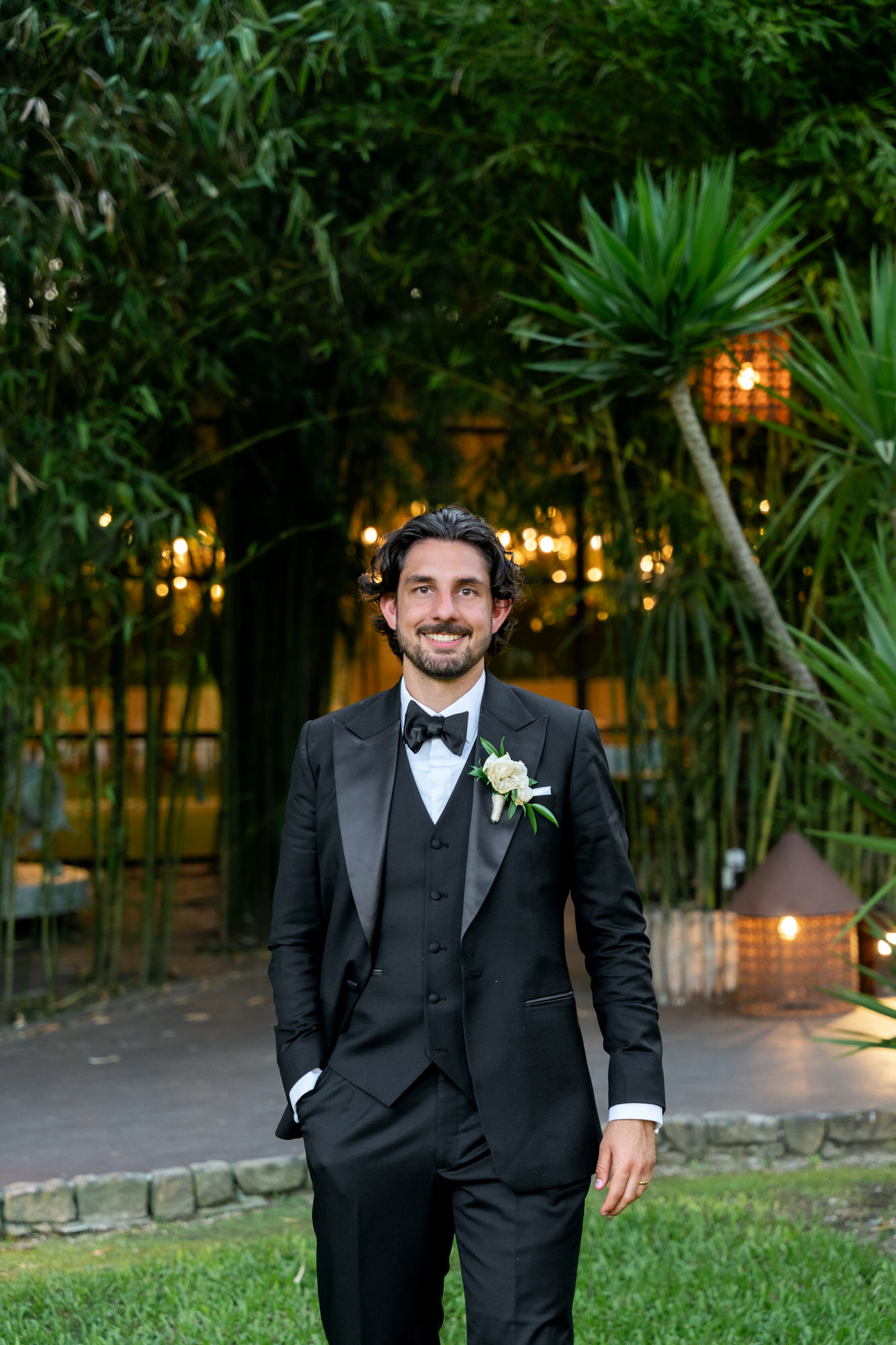 The groom in a black tuxedo and bow tie standing on the lawn, lanterns glowing behind him