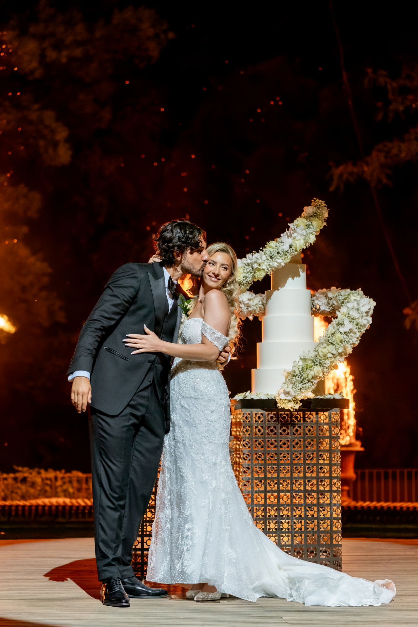 The couple kissing at night beside a stage of flames, a guest holding a bouquet behind them