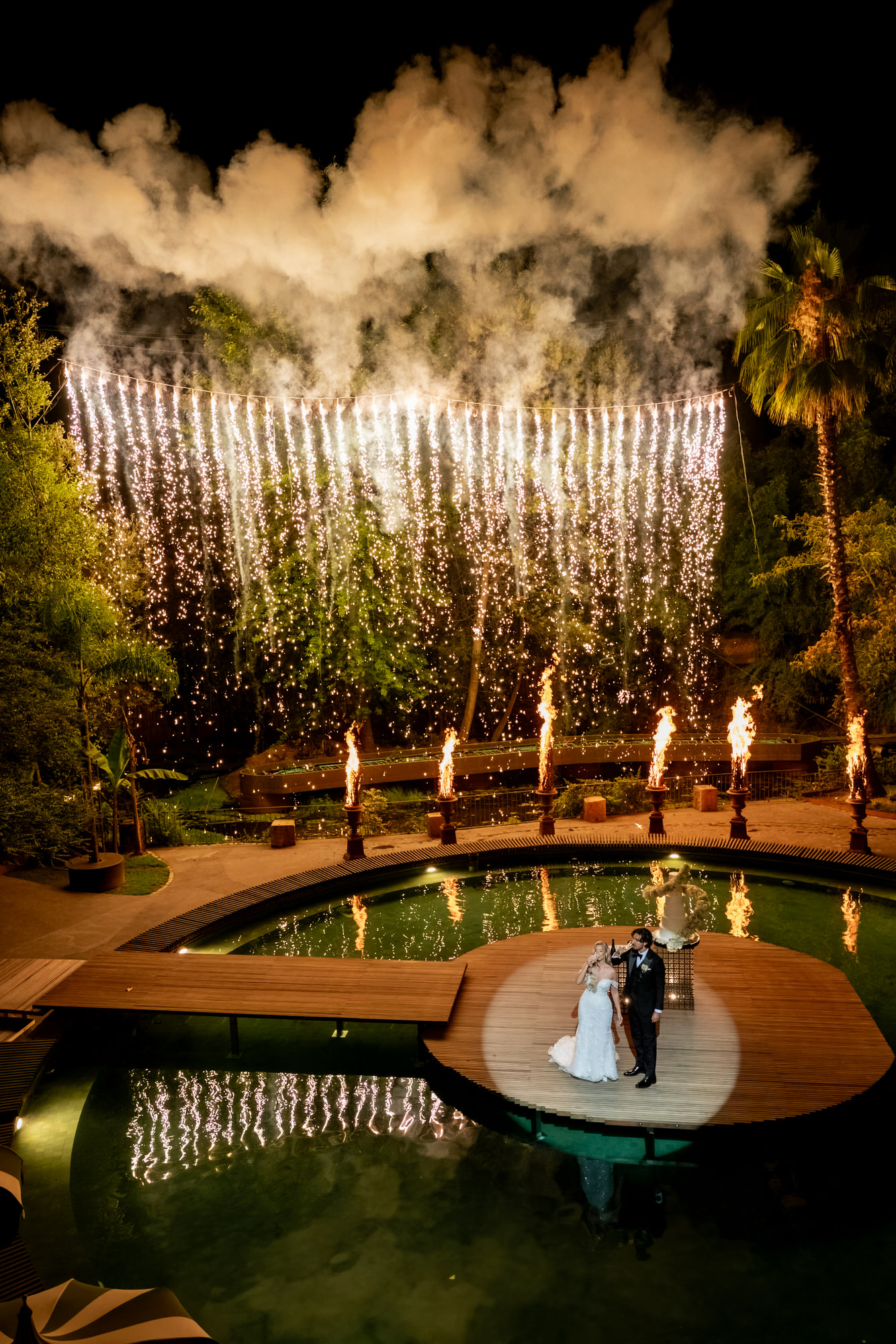 Wide night view of the couple on the lake platform, with floating candles and a curtain of sparks