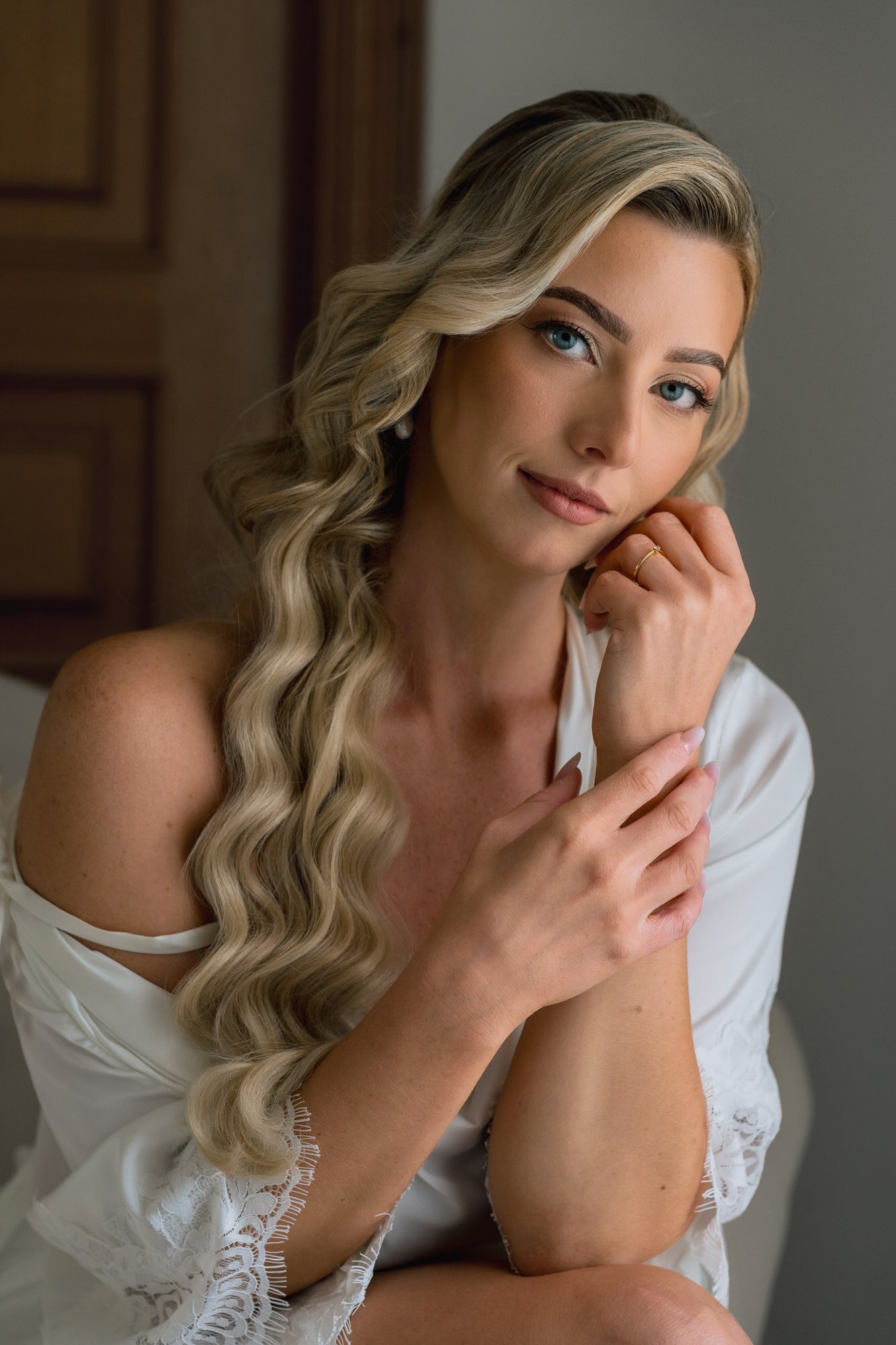 The bride in a white robe with long blonde waves, resting her chin on her hand