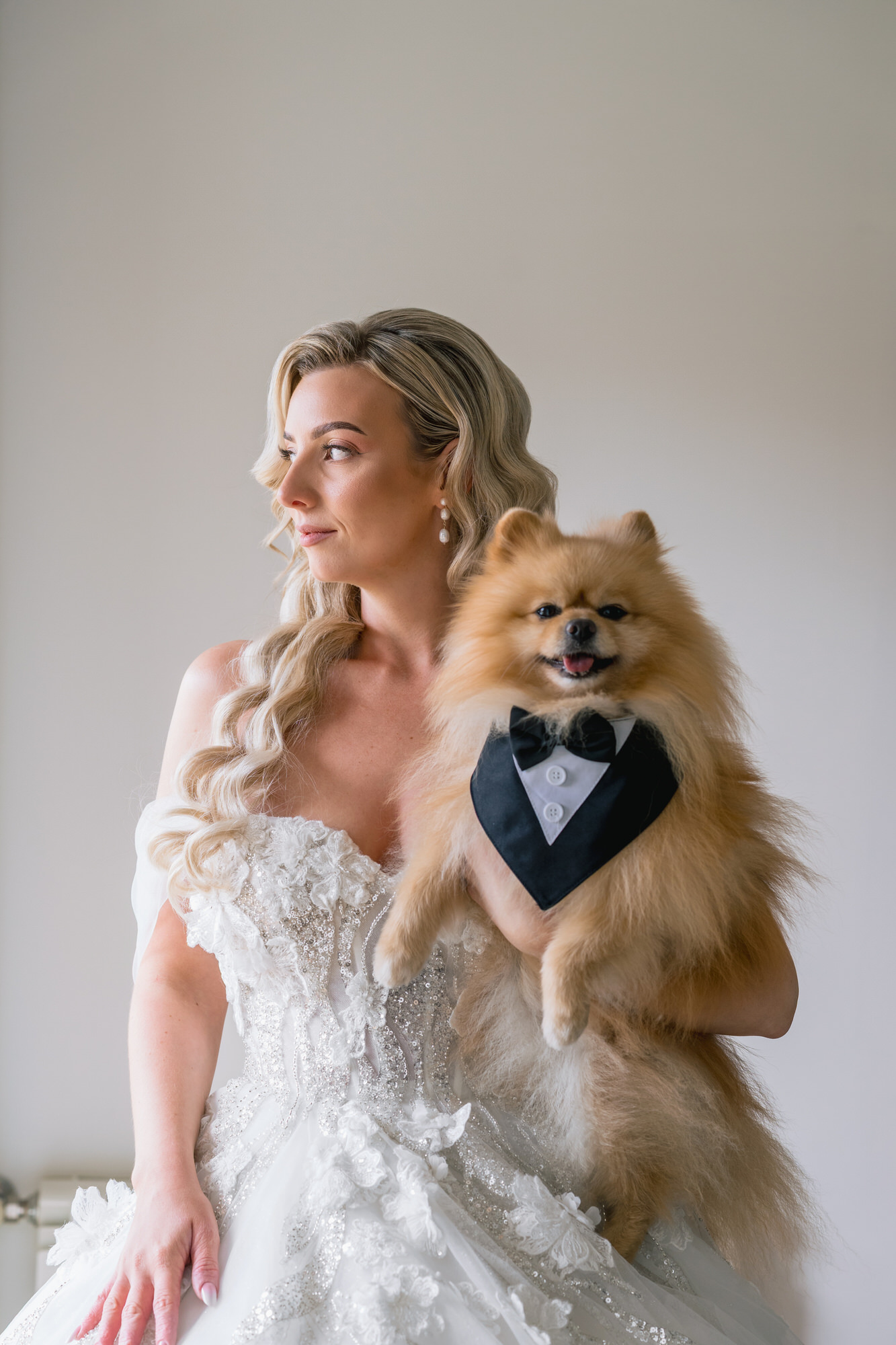 The bride in a lace gown holding her dog, who wears a tiny black bow tie