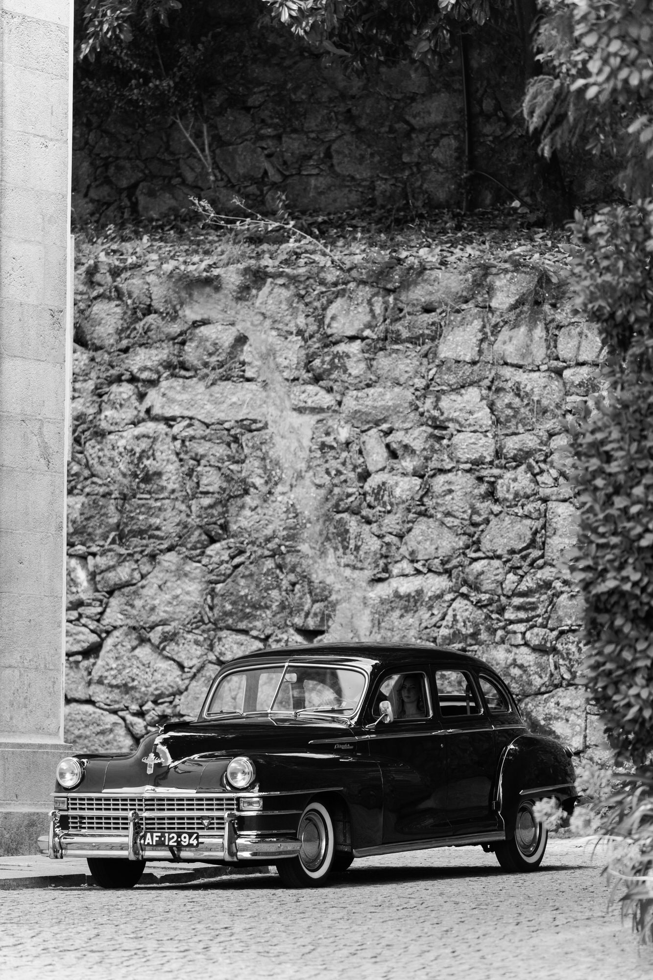 Black-and-white image of a classic car parked beside an old stone wall