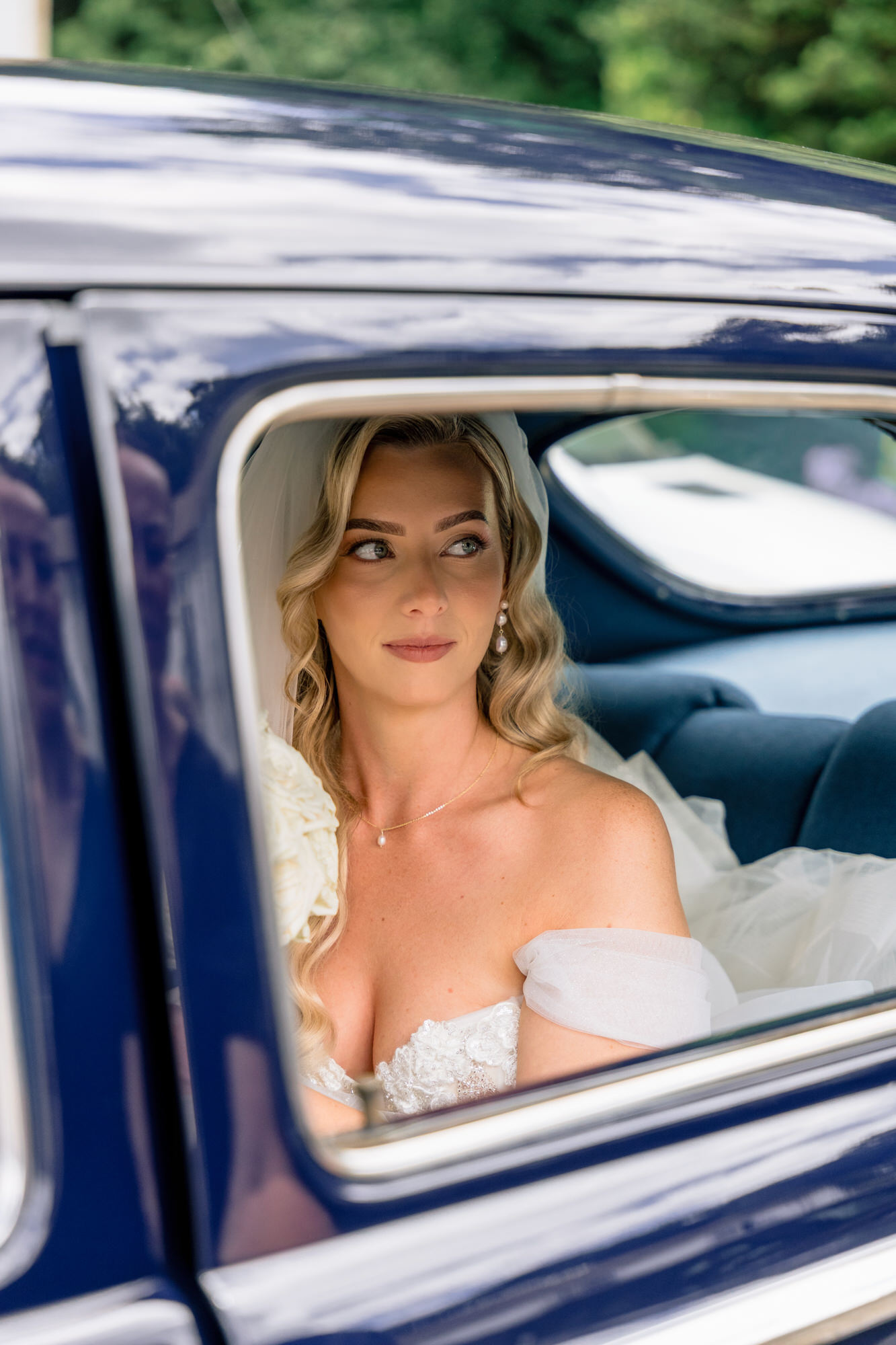 The bride in an off-the-shoulder gown looking out of the window of a navy vintage car