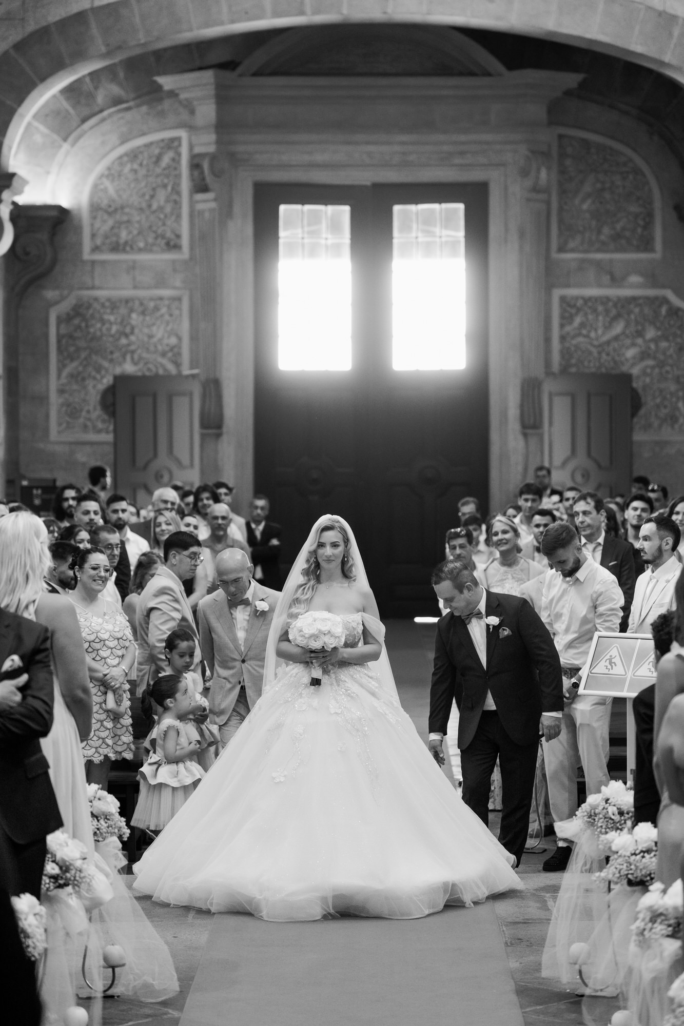 Black-and-white image of the bride in her ball gown entering the church, guests and flower girls around her