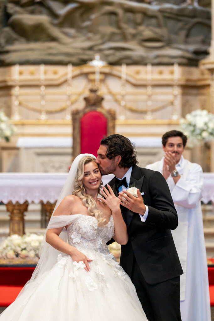 Bride and groom kiss at their wedding in a church, with a clapping guest in the background near an ornate altar.