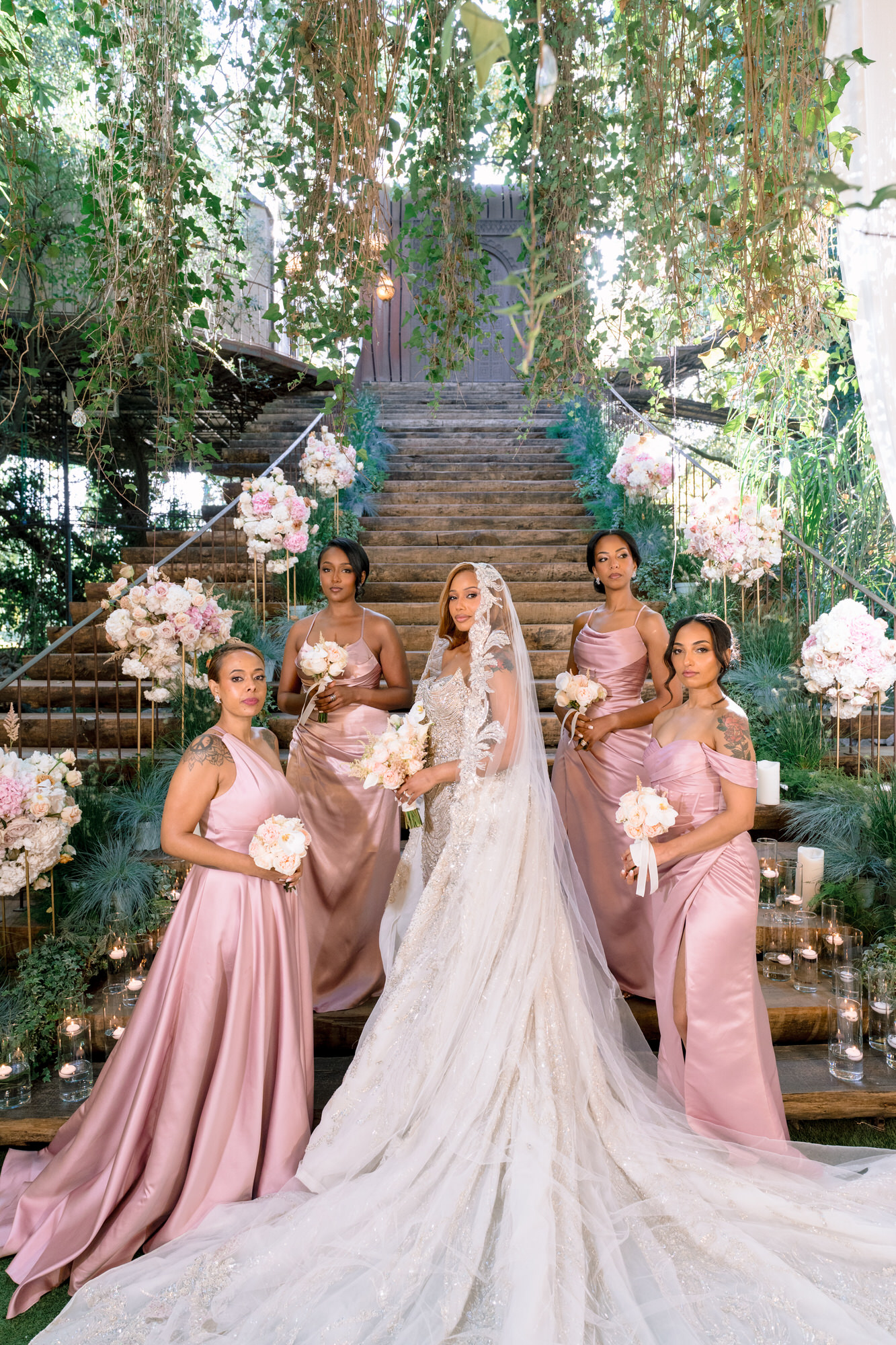 The bride with her bridesmaids in dusty pink dresses on the garden stairs, her train spread before them