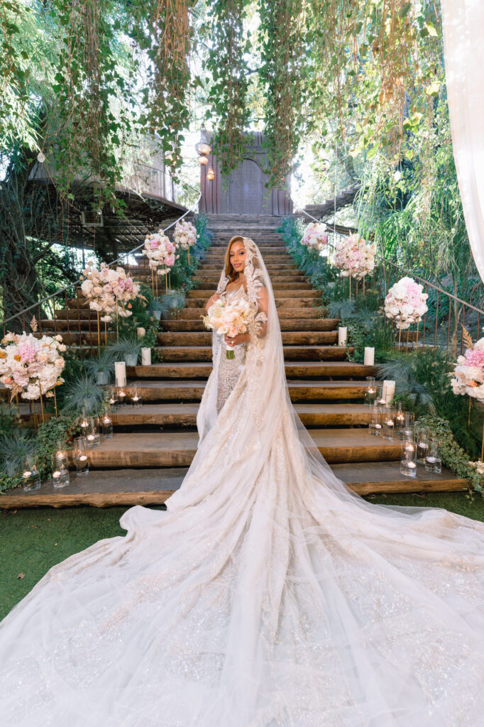 Bride in a white, glittering wedding gown and veil stands on a wooden staircase lined with pink and white flowers, holding a bouquet.