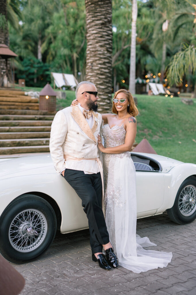 Couple in formal attire posing beside a white vintage car outdoors, both wearing sunglasses; man in cream suit and bow tie, woman in a beaded gown with a long tulle train, smiling at each other.