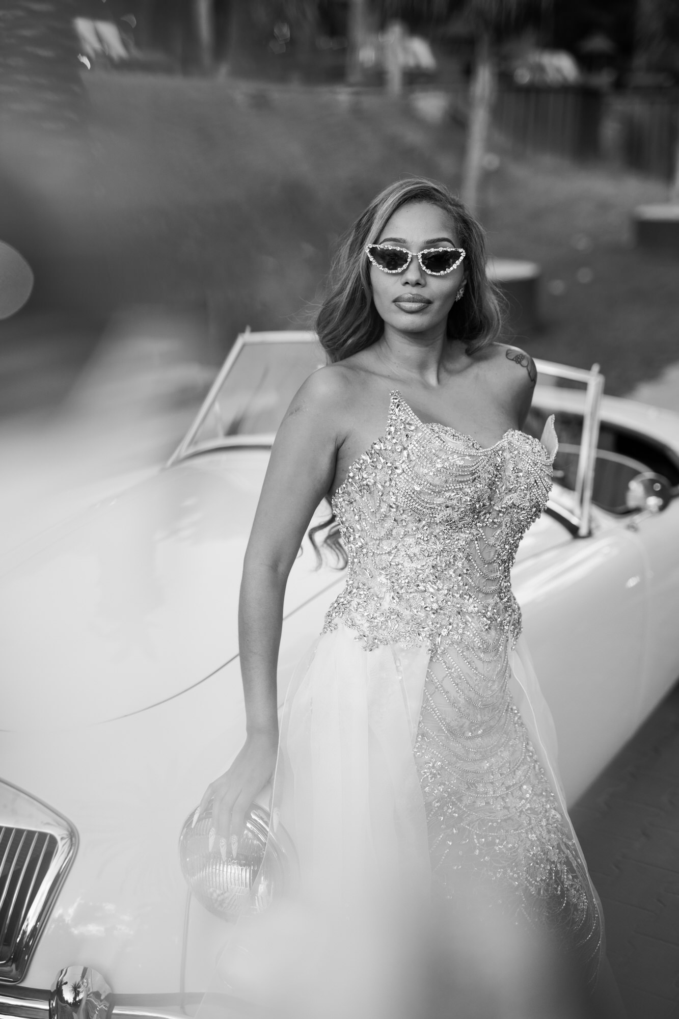 Black-and-white portrait of the bride in a sparkling gown and sunglasses beside the vintage car