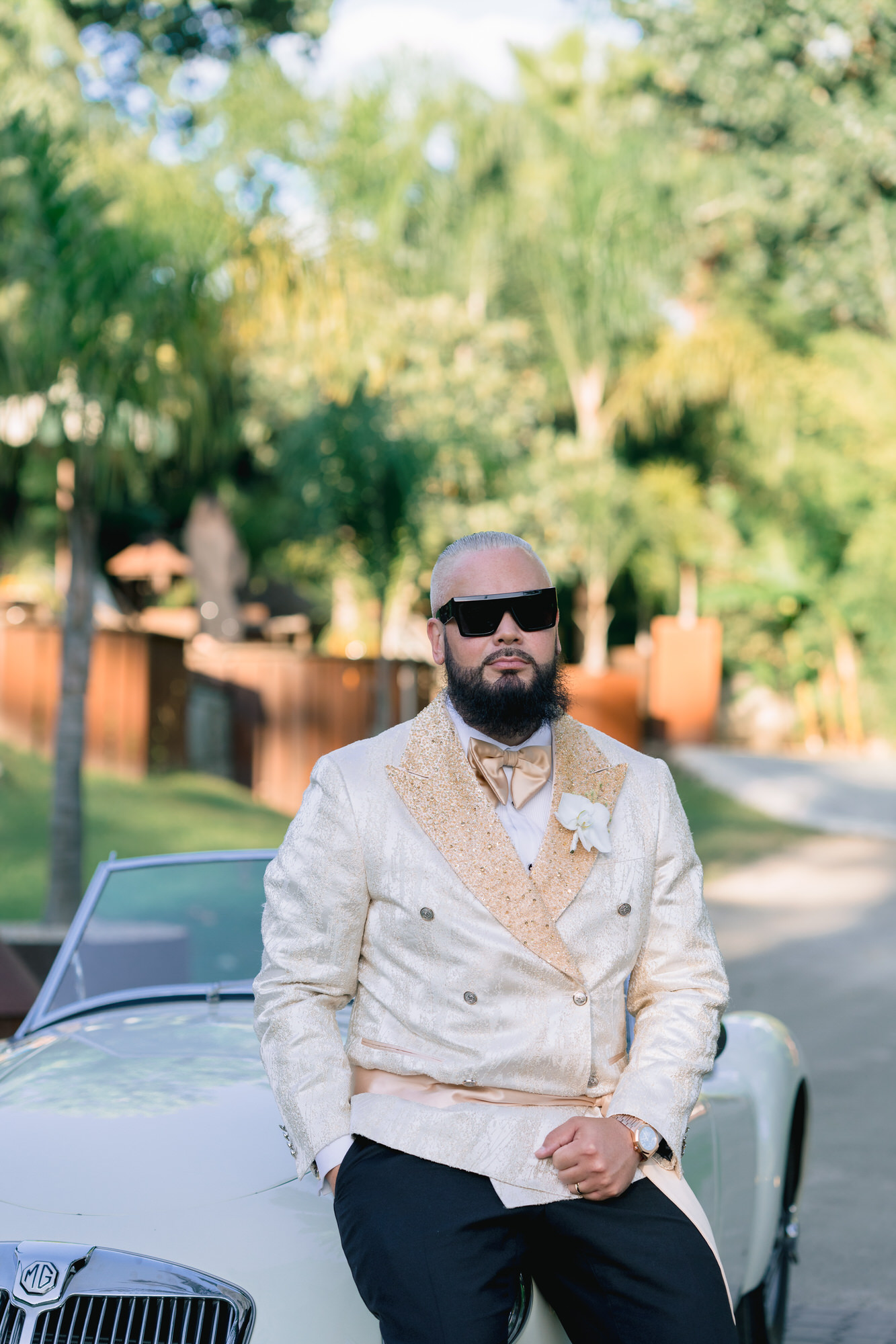 The groom in sunglasses and a cream double-breasted jacket leaning on a white vintage car