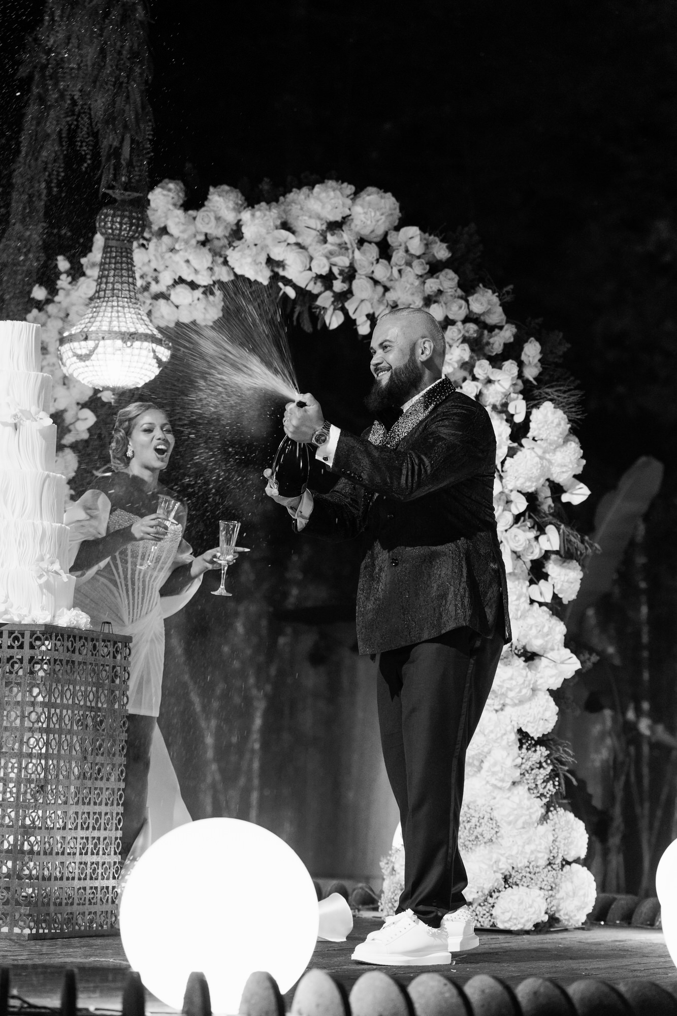 Black-and-white image of the couple at a white floral arch at night, a glowing orb in the foreground