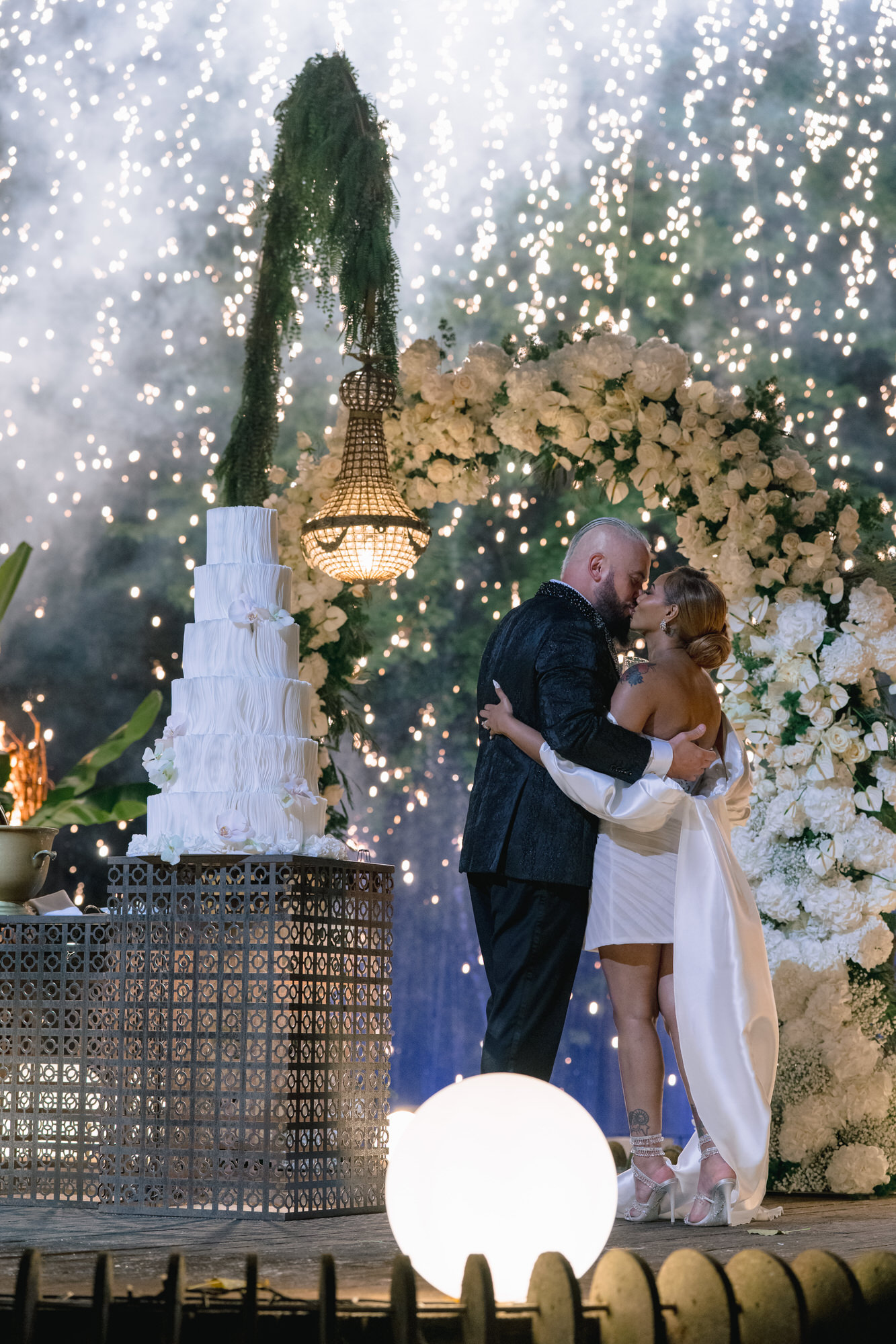 Mahlet and Thomas embracing beside a tall white cake in the woods of Quinta Lago dos Cisnes, fairy lights falling behind them