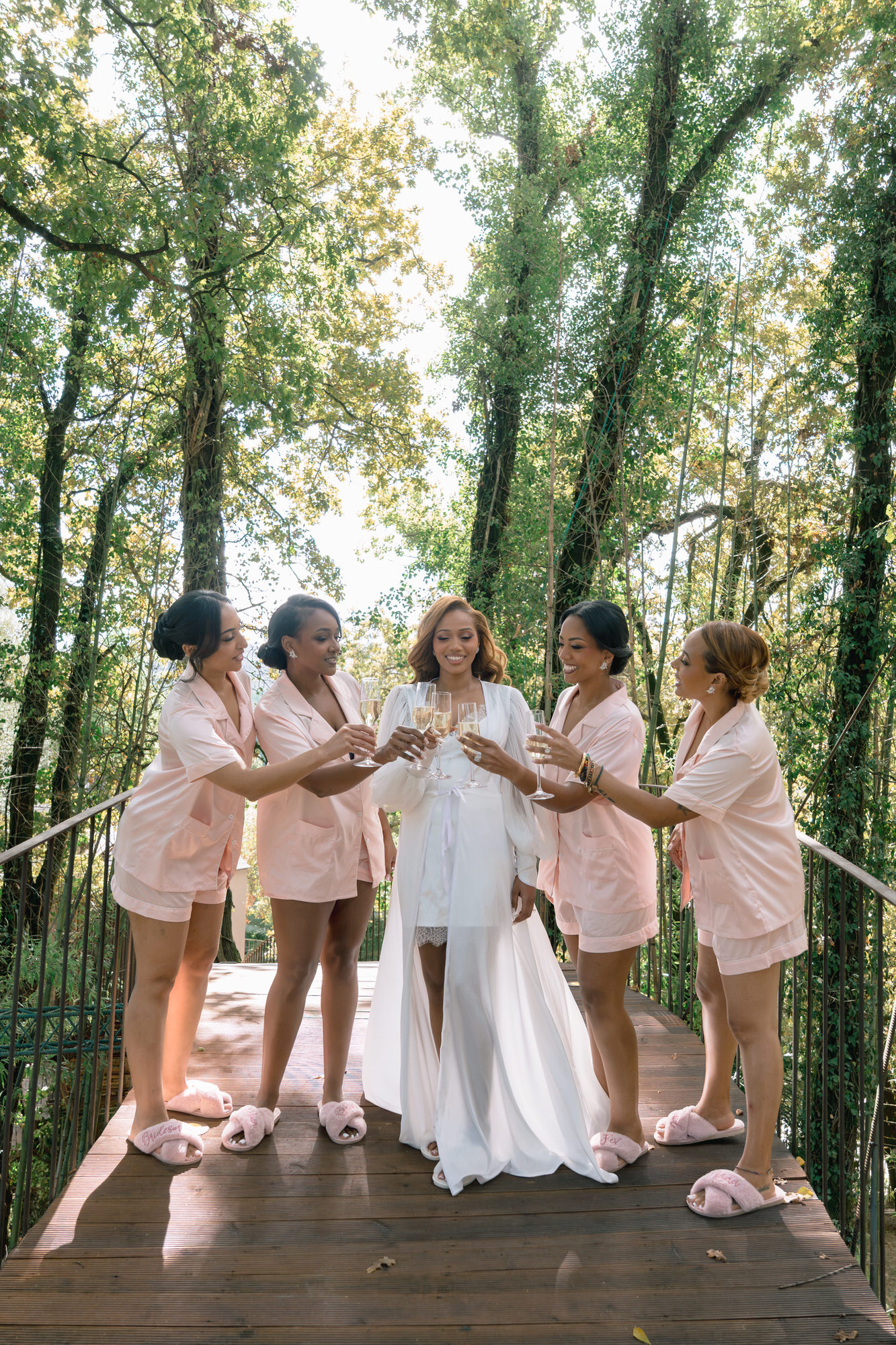 The bride in a white robe surrounded by her bridesmaids in blush robes on the forest boardwalk