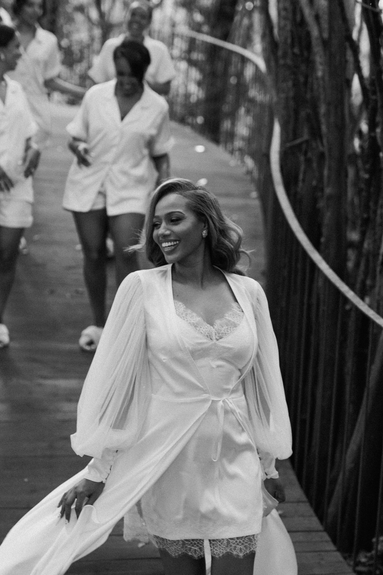 Black-and-white image of the bride in a flowing robe on the boardwalk, bridesmaids behind her