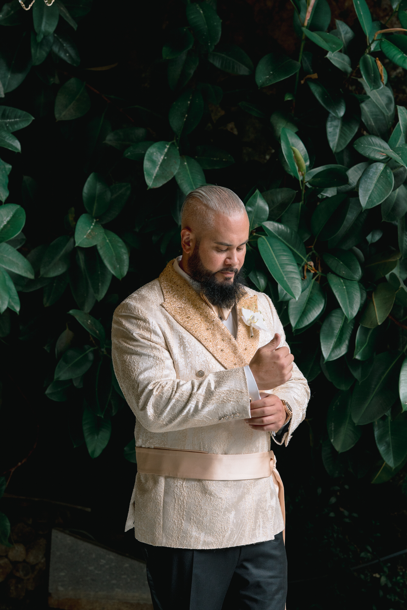 The groom in a cream jacket with gold trim, adjusting his cuff among dark green foliage