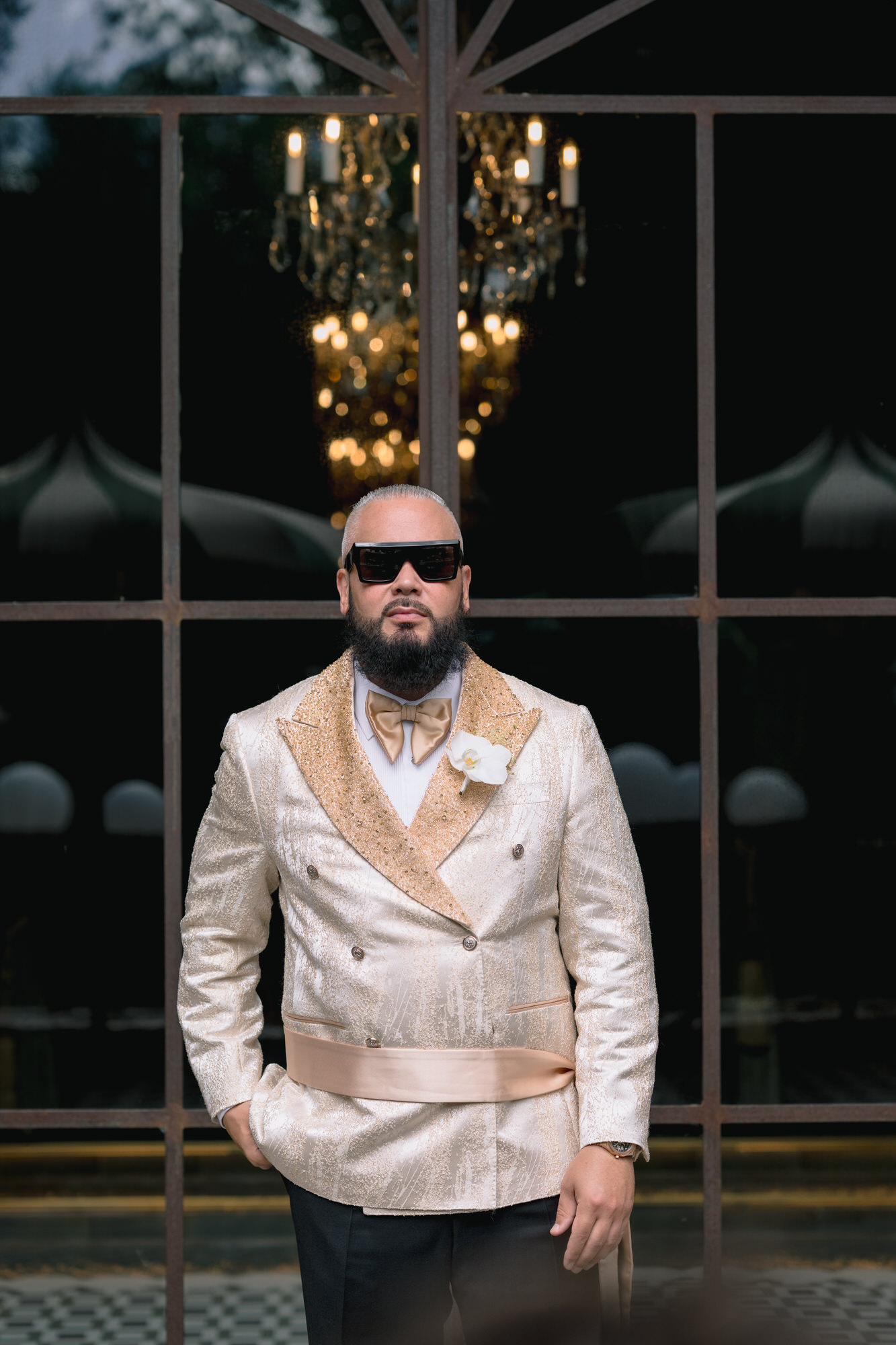 The groom in sunglasses and a cream jacket in front of dark glass doors and a chandelier