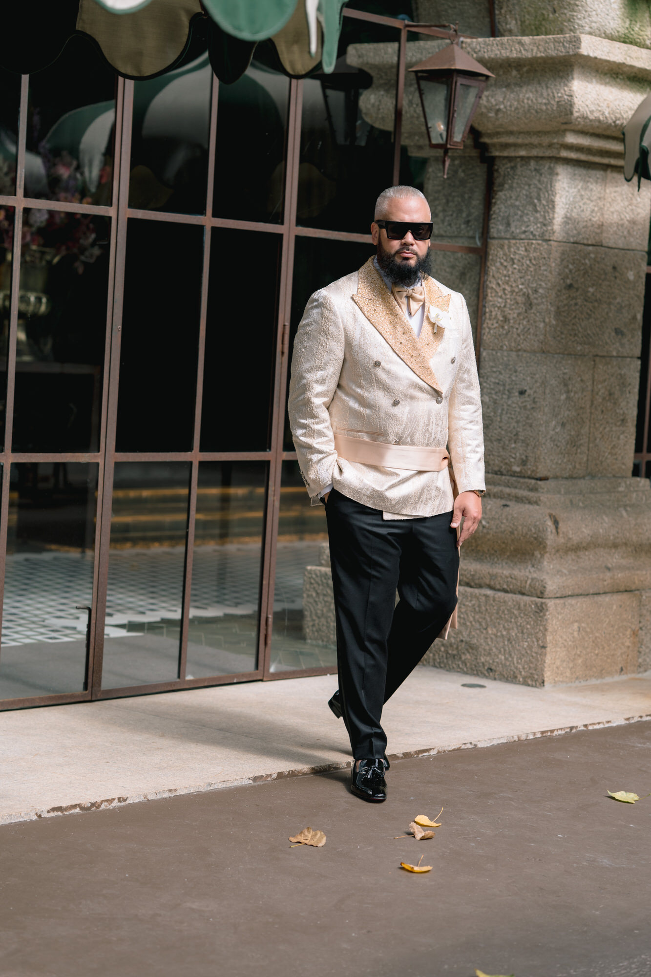 The groom in a cream dinner jacket walking along a stone wall beside tall glass windows